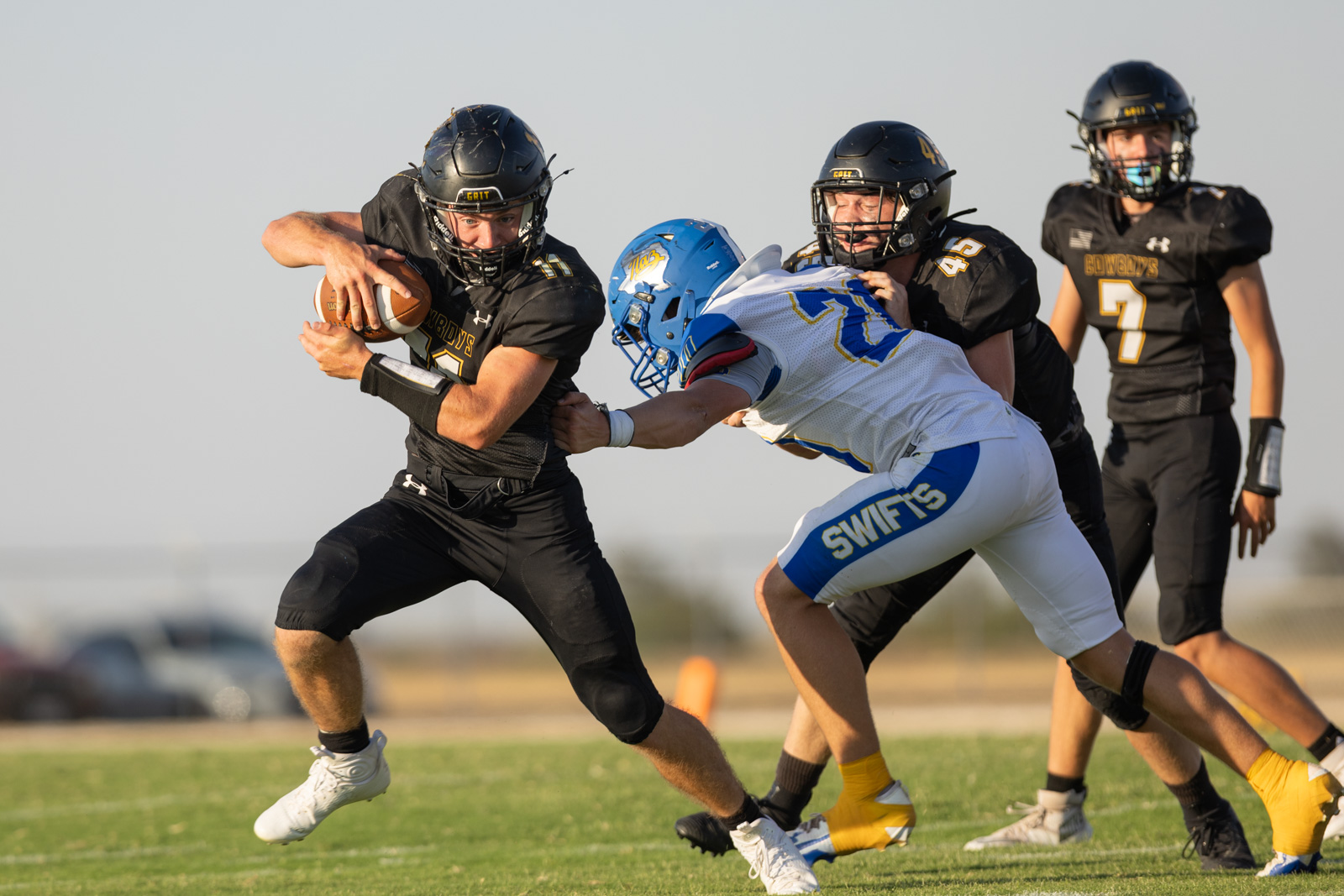 HHS-NHS-FB-30 | Press Pass Sports A football player in black jersey runs with the ball while a defender in white and blue tackles from the side on a grassy field.