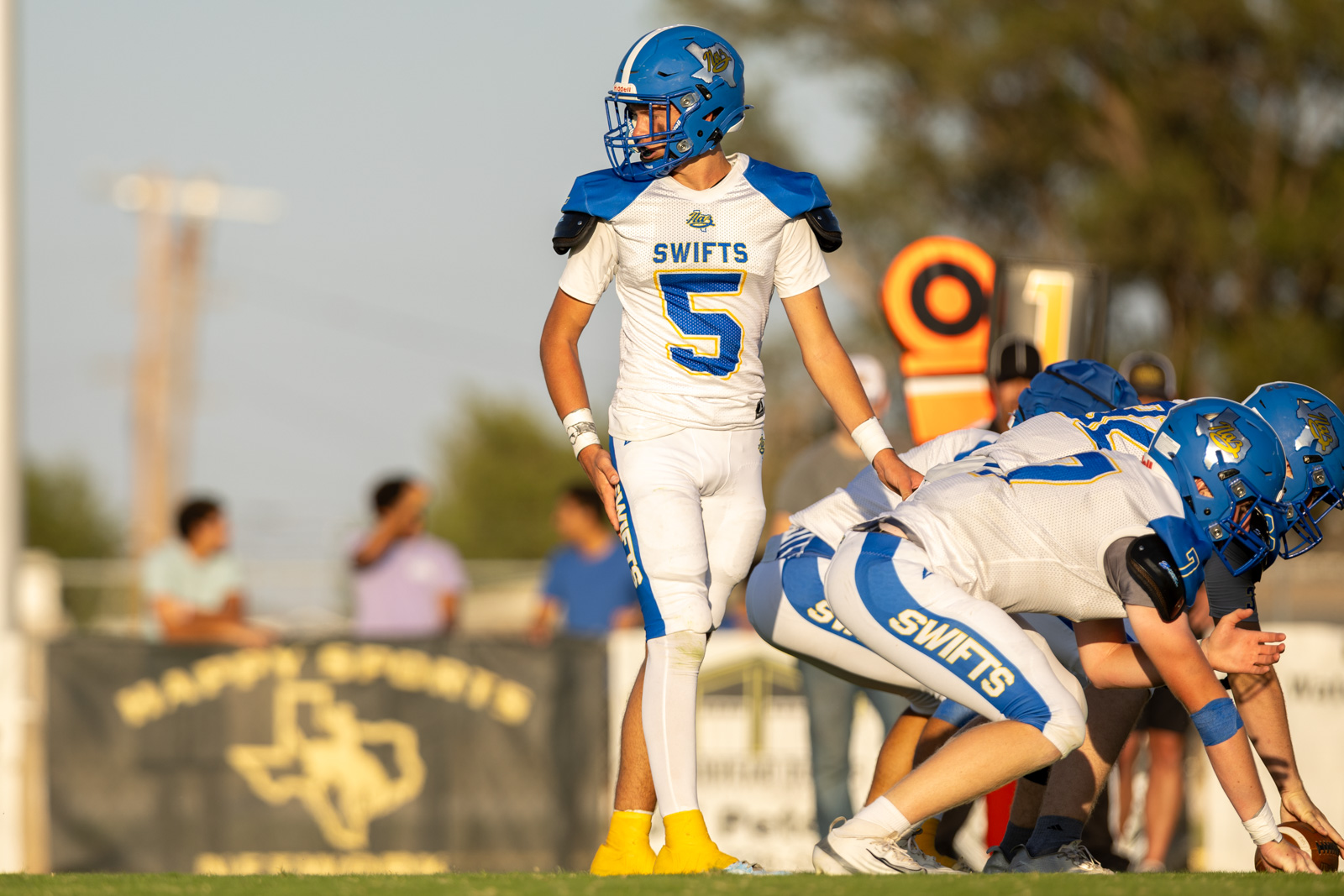 HHS-NHS-FB-31 | Press Pass Sports Young football player in white and blue uniform #5 stands over the line of scrimmage, teammates crouched behind him on the field.