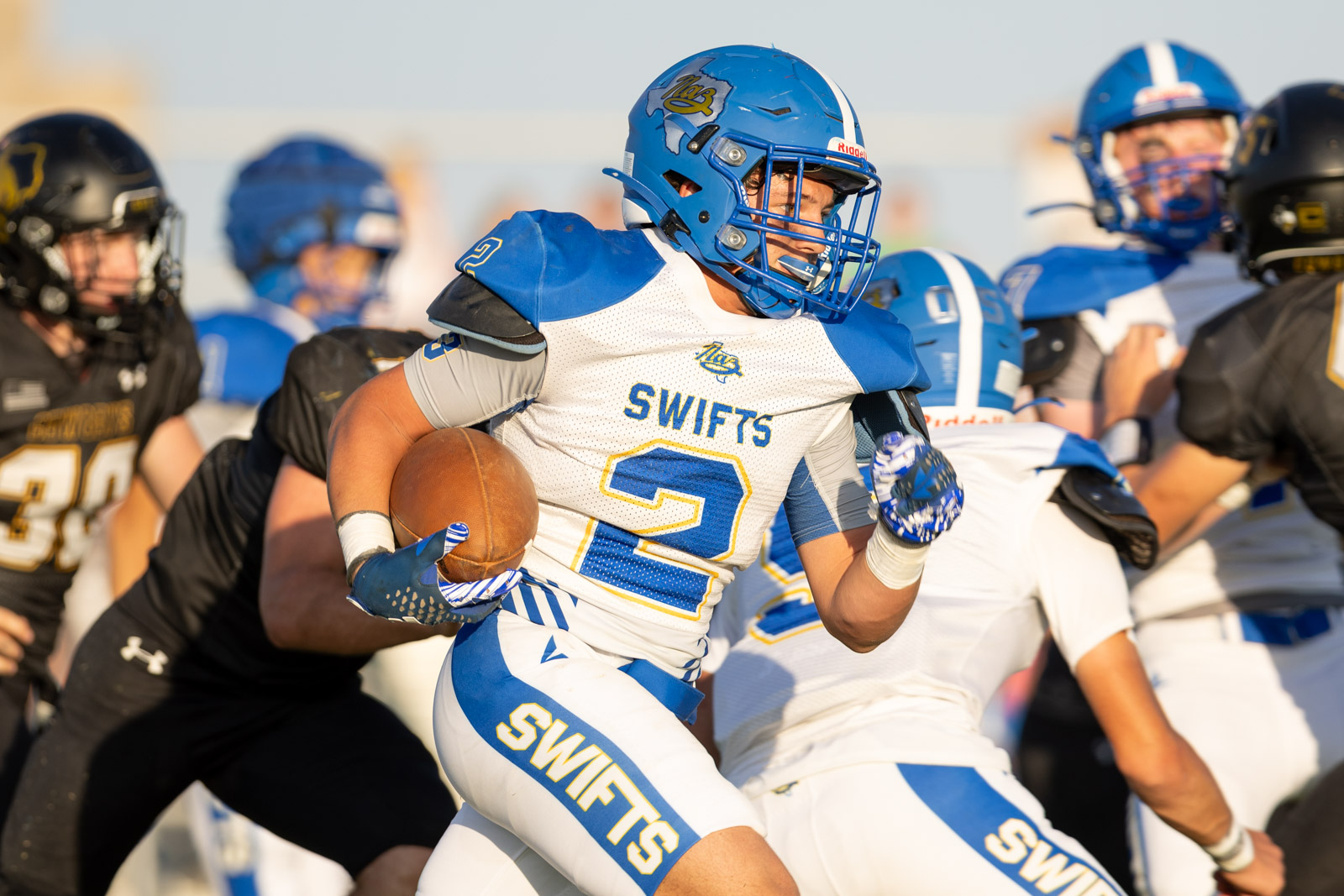 HHS-NHS-FB-33 | Press Pass Sports American football player in white and blue Swift uniform runs with the ball during a play, surrounded by defenders at a sunny field.