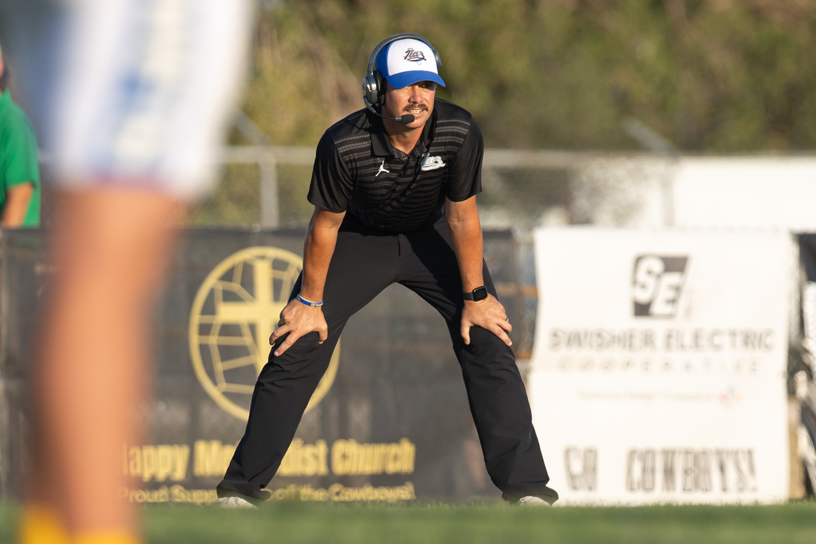 HHS-NHS-FB-34 | Press Pass Sports Coach wearing a headset crouches on the field, focused on the play ahead. The coach wears a black polo and blue-and-white cap.