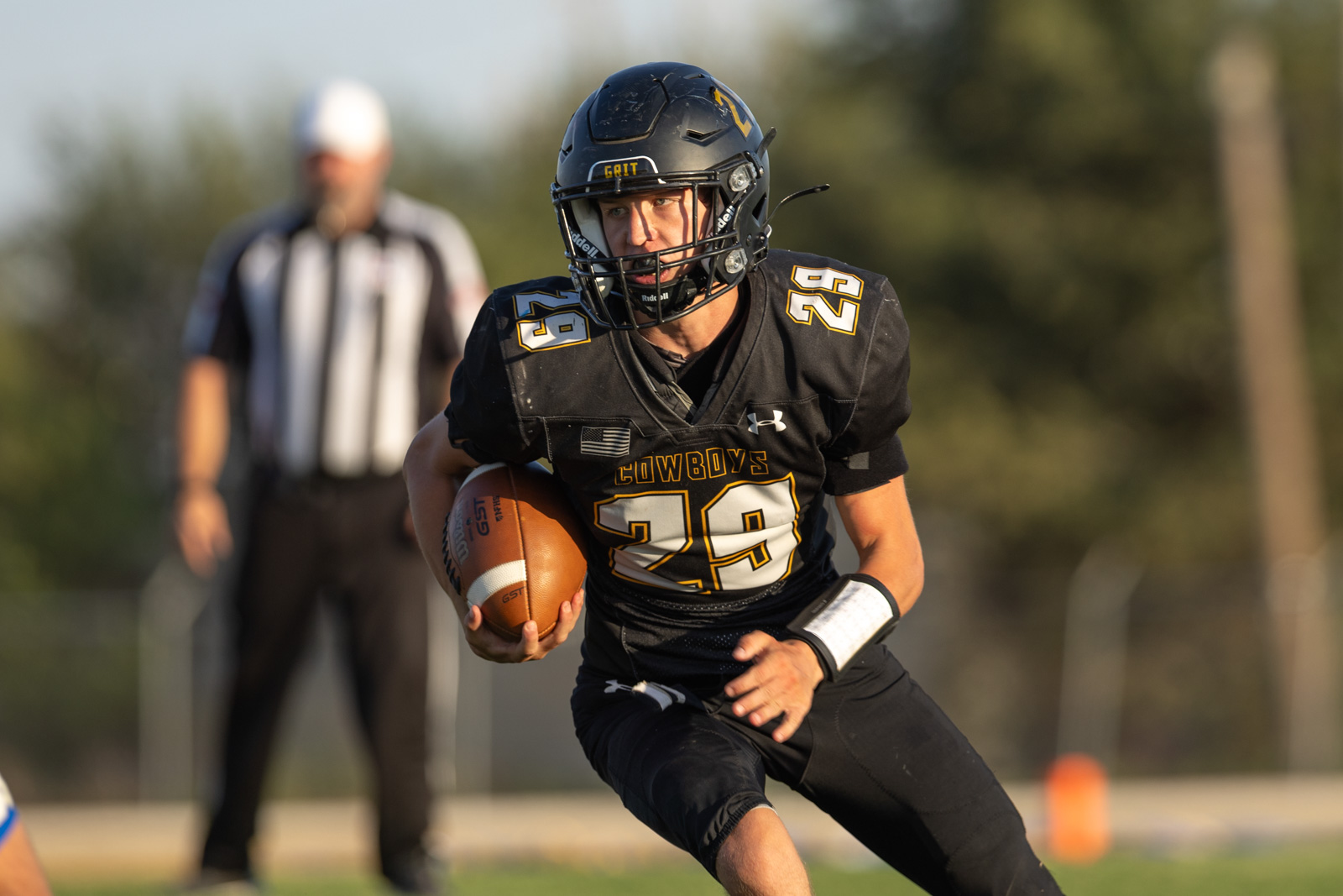 HHS-NHS-FB-35 | Press Pass Sports Young football player in a black Cowboys jersey holds the ball and runs forward on the field with a referee in the background.