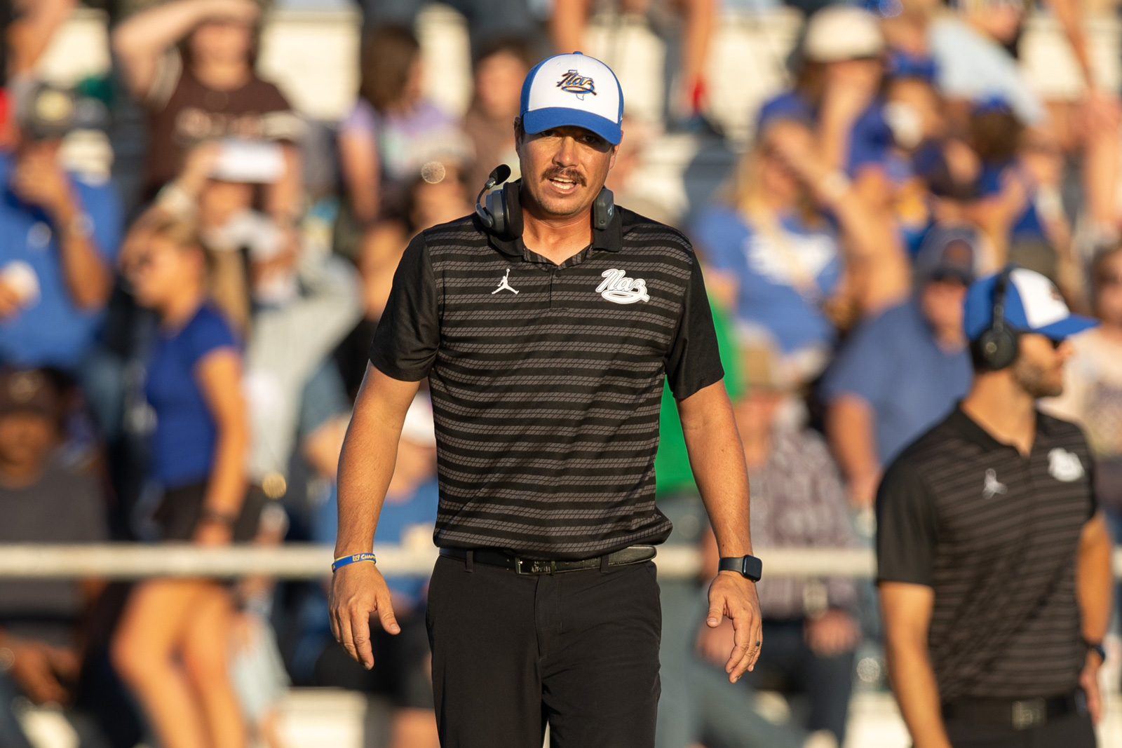 HHS-NHS-FB-36 | Press Pass Sports Man wearing a black striped polo and cap with headset around his neck walks toward camera at a crowded sports venue.