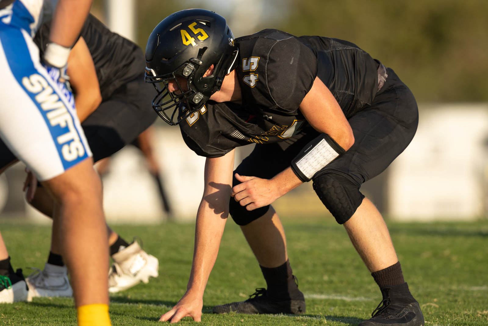 HHS-NHS-FB-37 | Press Pass Sports American football lineman in a black uniform crouches at the line of scrimmage, helmet number 45, ready for the snap on a sunny field.