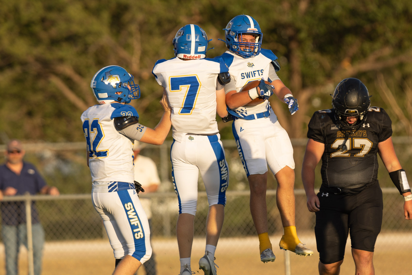 HHS-NHS-FB-38 | Press Pass Sports Three football players in white and blue uniforms jump mid-air in celebration on the field as teammates hug, while a dark-colored opposing player watches nearby.