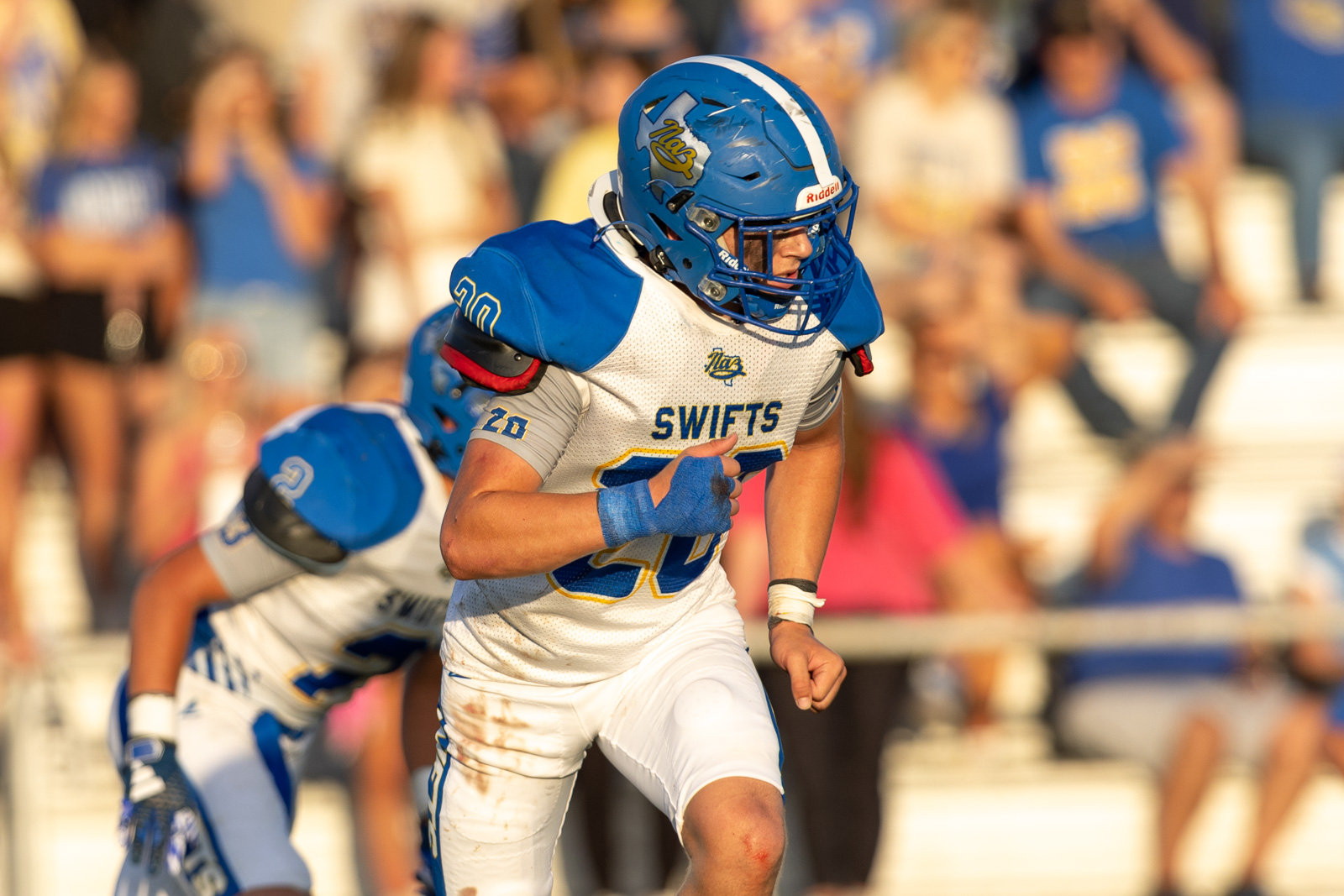 HHS-NHS-FB-40 | Press Pass Sports Football player in blue helmet and white-and-blue uniform runs forward with the ball during a game, crowd blurred in the stands behind.