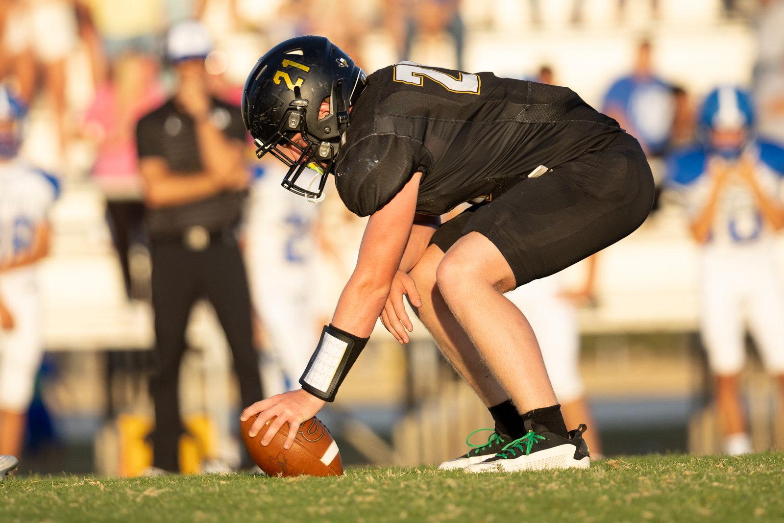 HHS-NHS-FB-41 | Press Pass Sports Football center in black uniform crouches over the ball, preparing to snap on a sunlit field with blurred crowd in the background