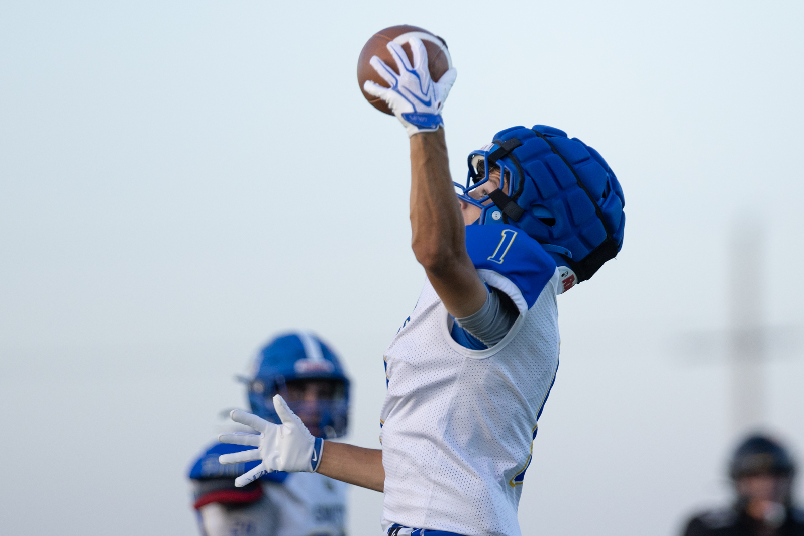HHS-NHS-FB-48 | Press Pass Sports Football player leaps to catch a ball in midair, wearing a white jersey with blue trim and a blue helmet with gloves on.