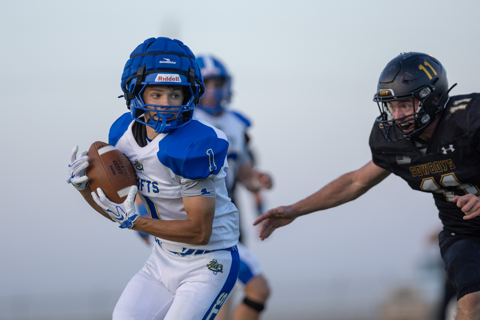 HHS-NHS-FB-49 | Press Pass Sports Football action: a blue-and-white team player runs with the ball, defender in black pursuing close behind.