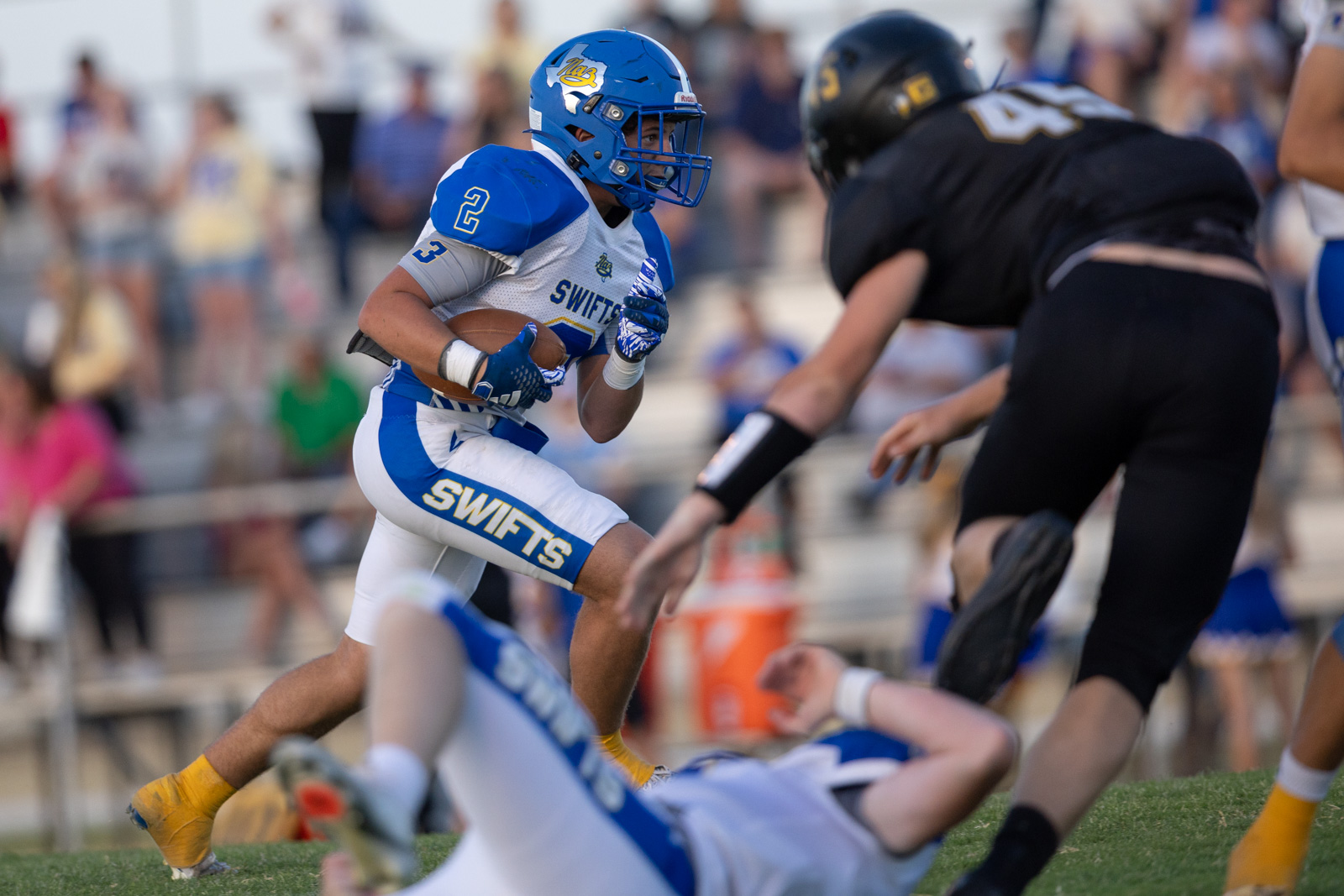 HHS-NHS-FB-50 | Press Pass Sports Youth football player in blue and white SWIFTS uniform runs with the ball as defenders approach nearby crowds blur in the background.