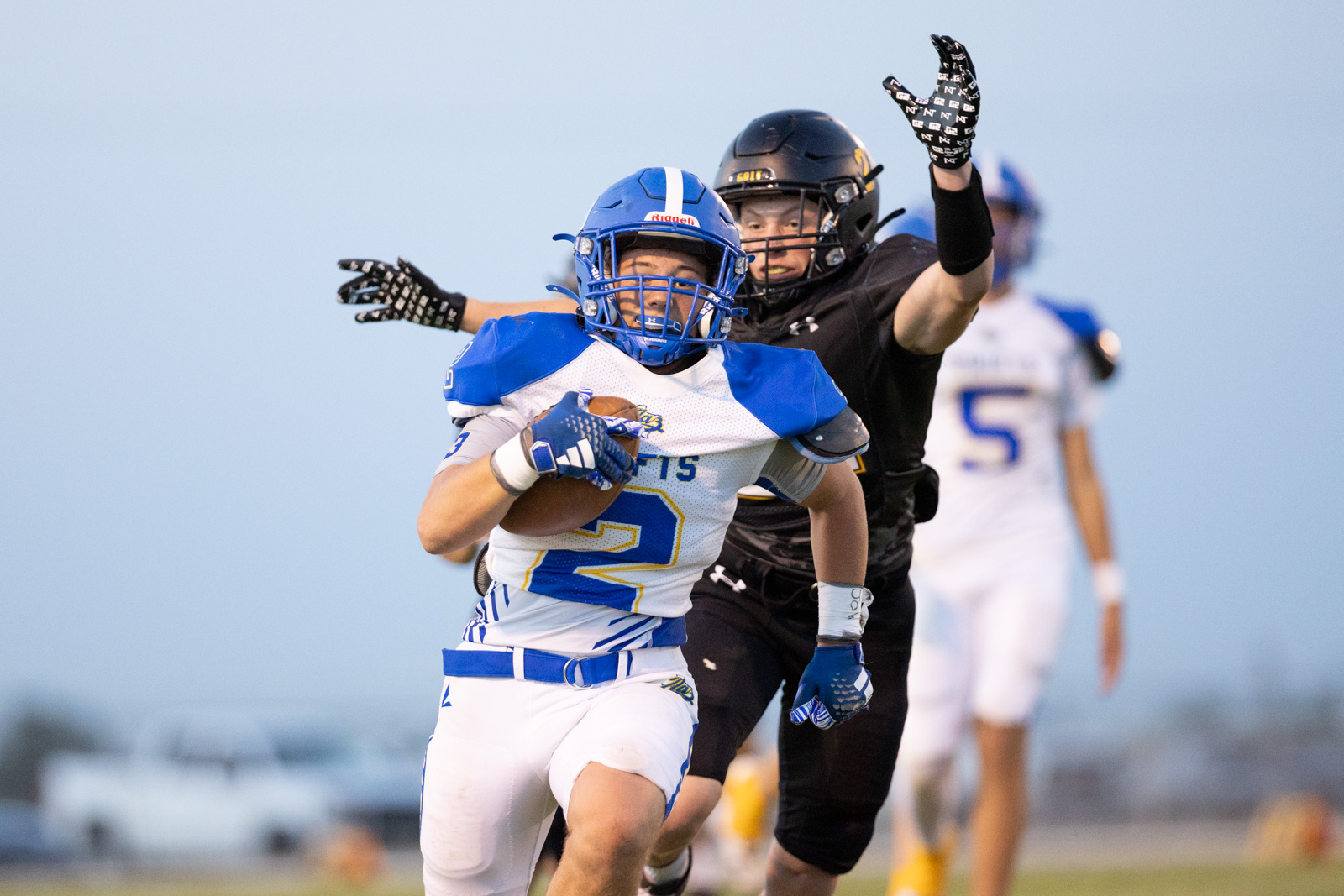 HHS-NHS-FB-52 | Press Pass Sports Football player in blue and white uniform runs with ball, sprinting ahead while a defender in black reaches from behind as the play unfolds.