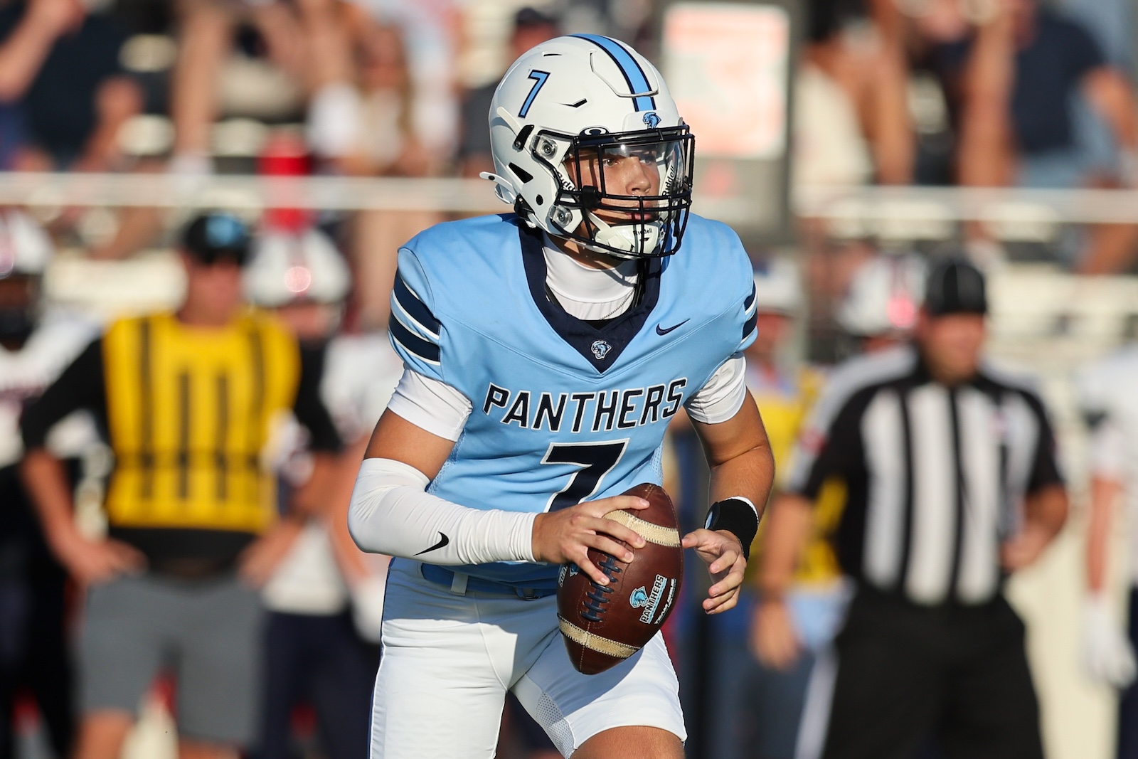 Seabolt Chase-14169 | Press Pass Sports Panthers quarterback in a light-blue jersey with number 7 holds a football during a game on the field with a referee in the background.