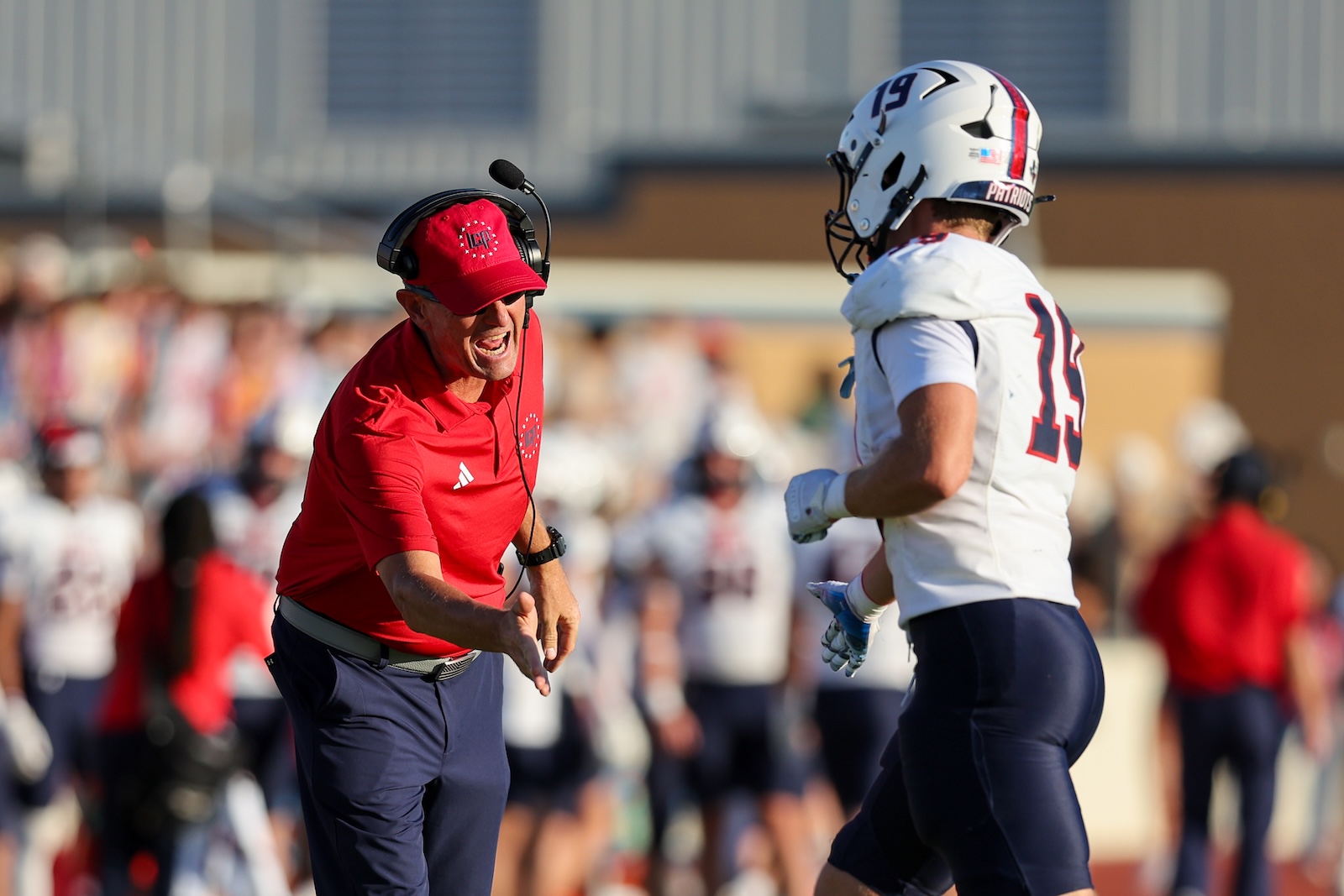 Seabolt Chase-14245 | Press Pass Sports Football coach in red shirt and headset shouting at a player in white with a Patriot helmet during a game. Crowd blurred in the background.