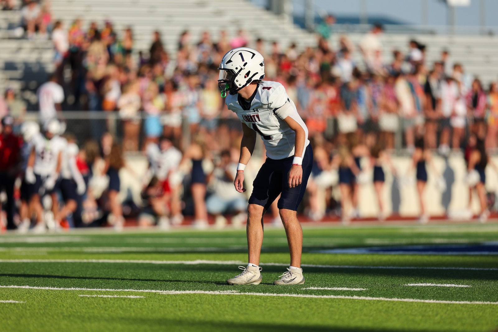 Seabolt Chase-14285 | Press Pass Sports Football player in white jersey and helmet crouches on a green field with a crowded stadium in the background, ready for play.