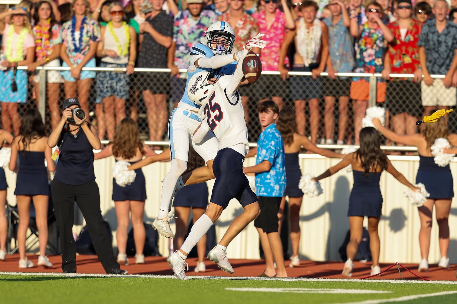 Seabolt Chase-14394 | Press Pass Sports Two football players leap for a catches mid-air as a pass is contested, with a cheering crowd behind them on the stands.”