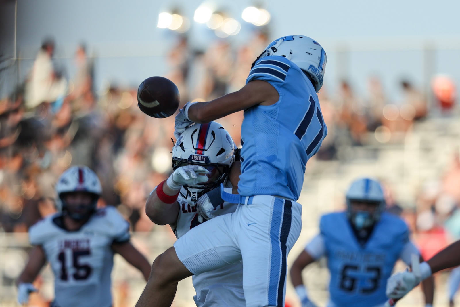Seabolt Chase-14441 | Press Pass Sports Two football players in light blue uniforms jump for a loose ball during a game, with a blurred crowd in the background.
