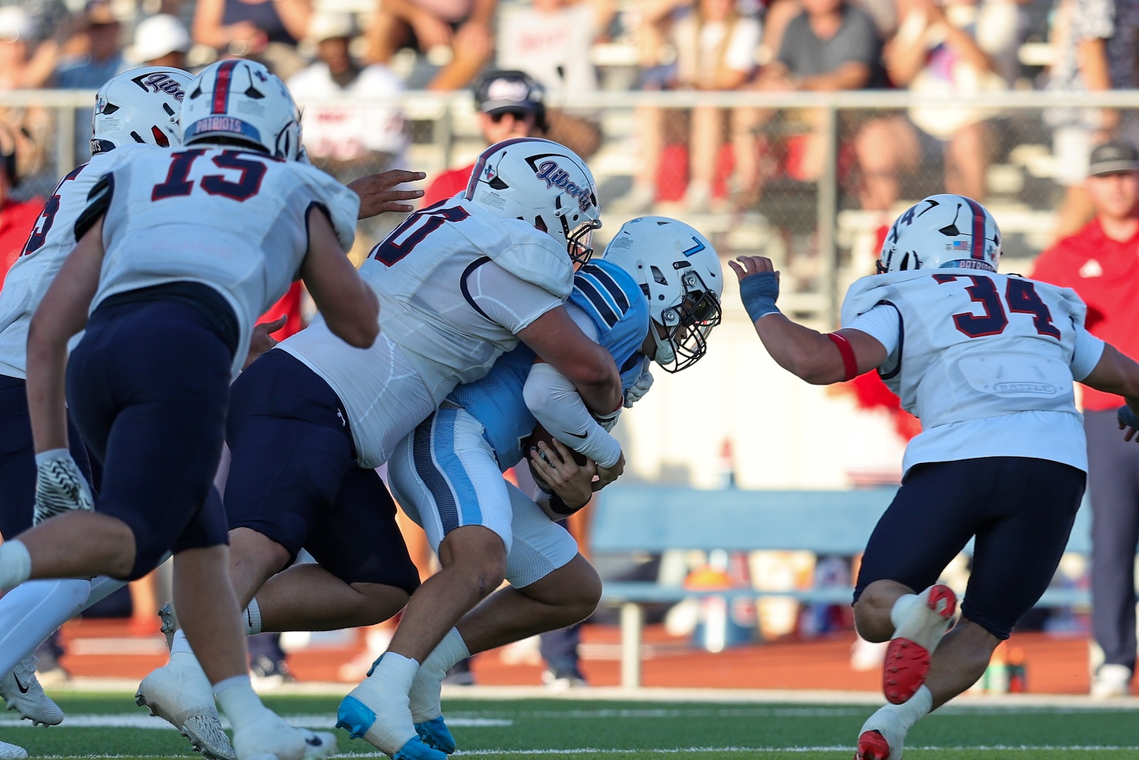 Seabolt Chase-14477 | Press Pass Sports Football players in white and navy tackle a ball carrier in light blue during a game on a sunny day.