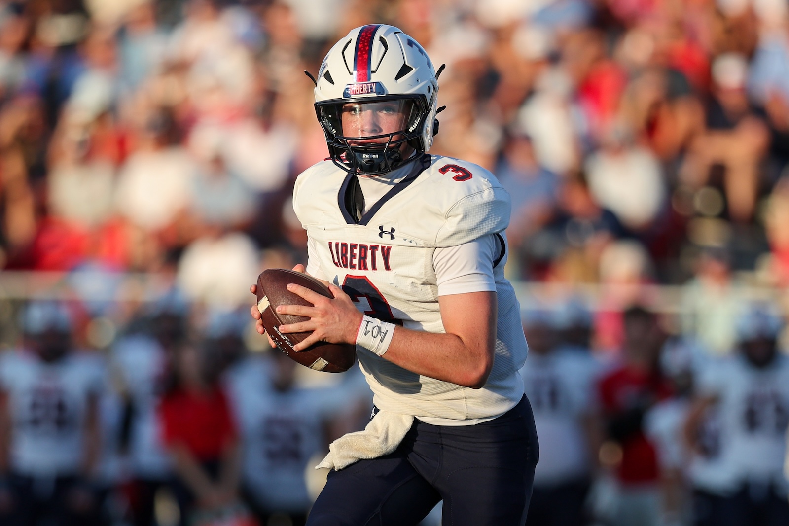 Seabolt Chase-14677 | Press Pass Sports A Liberty football quarterback holding a football, running on the field with a crowd in the background.