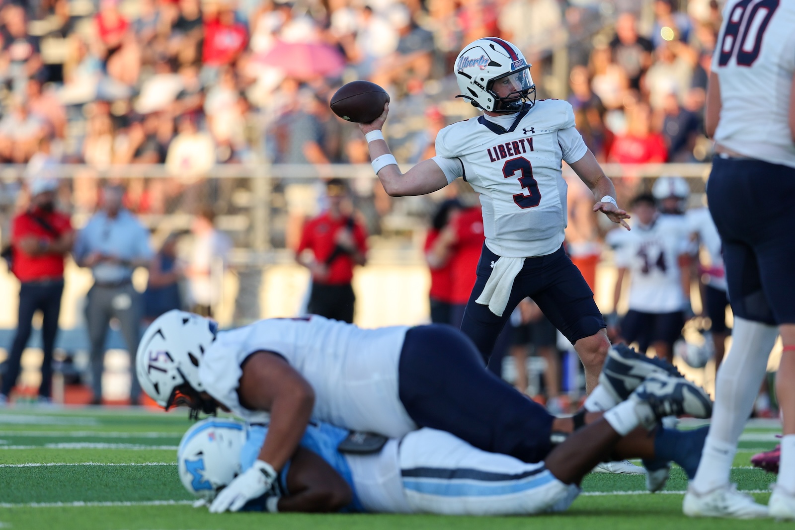 Seabolt Chase-14970 | Press Pass Sports Quarterback in a Liberty white jersey (number 3) throwing a pass while teammates and opponents collide on the field, crowd in the stands behind.