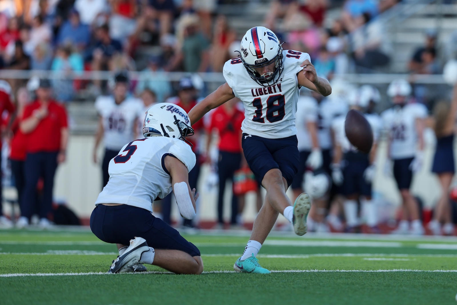 Seabolt Chase-15027 | Press Pass Sports Liberty football player in white #18 jumps to kick the ball while a defender crouches nearby on the field as the crowd watches in the stands.