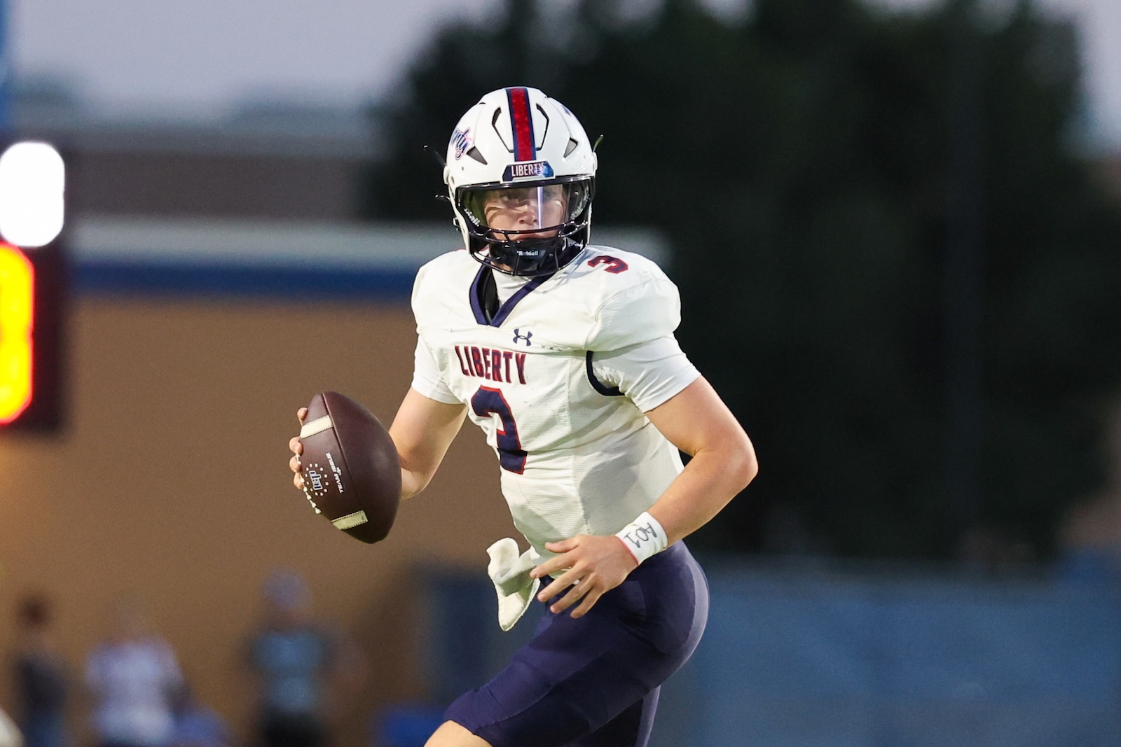 Seabolt Chase-15185 | Press Pass Sports Liberty football quarterback in a white jersey holding a football and running on the field, number 3 visible.