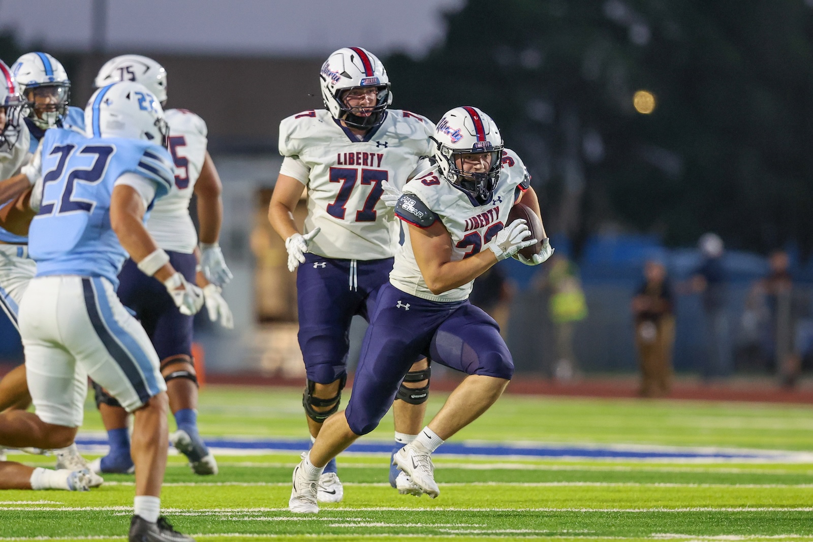 Seabolt Chase-15214 | Press Pass Sports Liberty football player in white jersey carries the ball while teammates block on a bright green turf field at dusk/dawn gymnastics-style lighting.