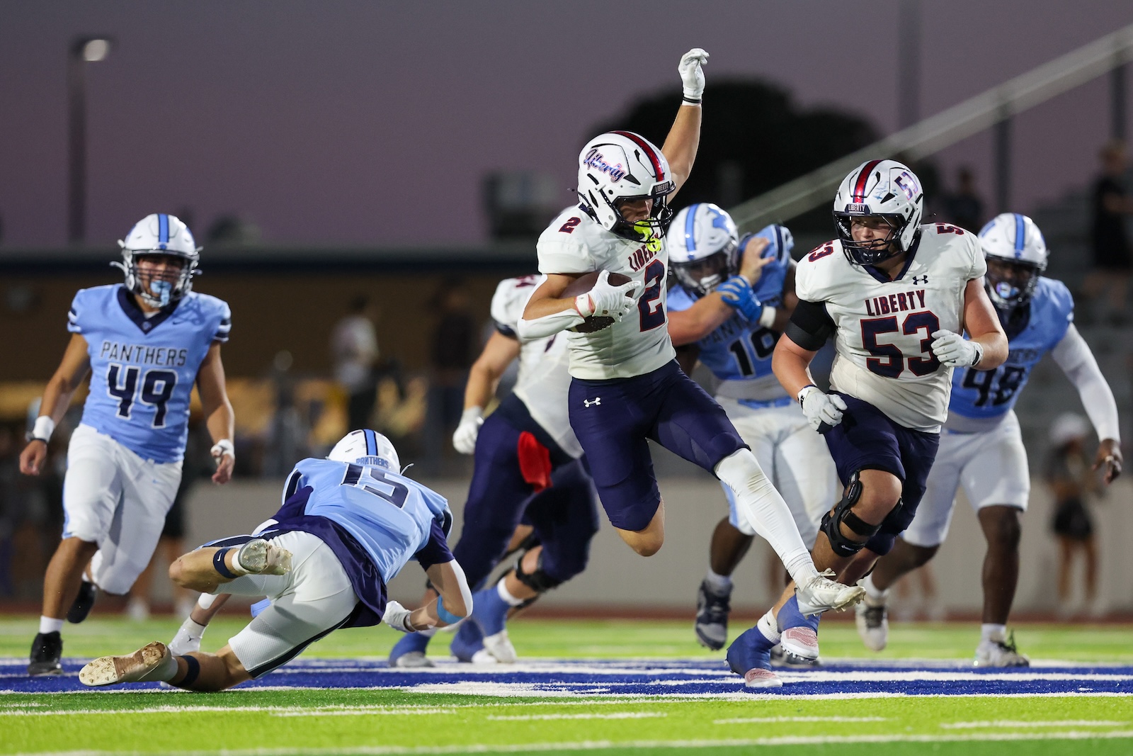 Seabolt Chase-15552 | Press Pass Sports Liberty players in white jerseys chase a ball carrier on a brightly lit football field during a night game with Panthers defenders close behind.