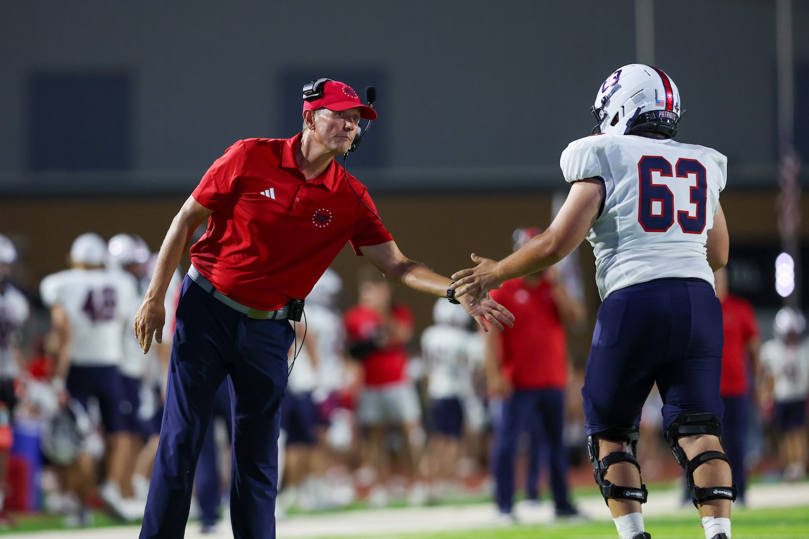 Seabolt Chase-15656 | Press Pass Sports Football coach in red polo and headset shaking hands with a football player wearing number 63 on the sideline during practice or a game.