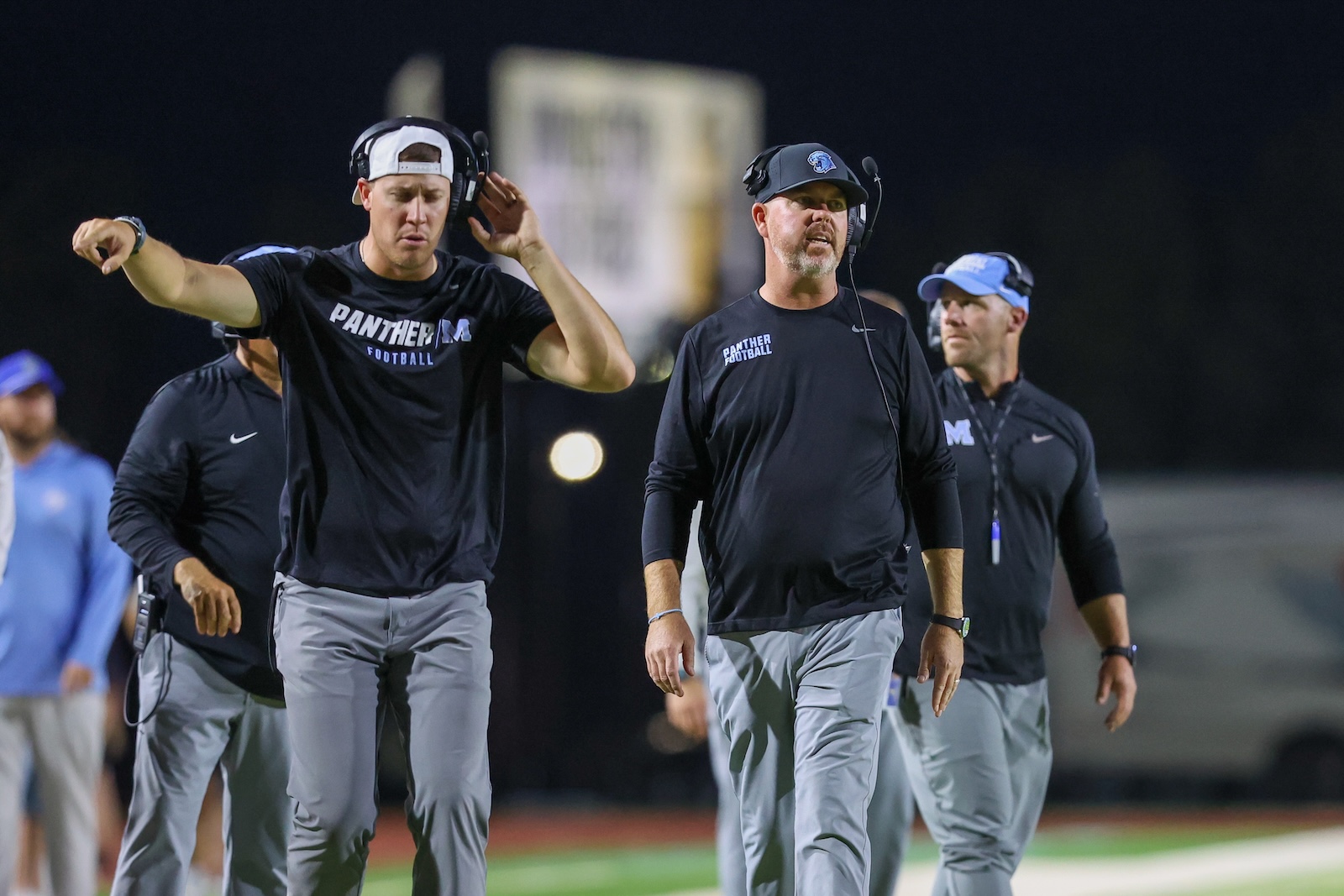 Seabolt Chase-16046 | Press Pass Sports Coaches in black Panthers shirts walking on a football field at night, wearing headsets and listening.