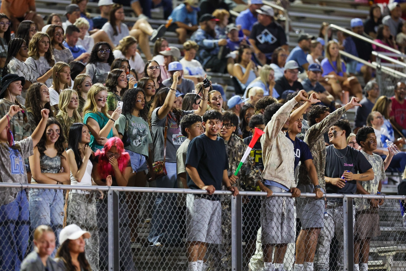 Seabolt Chase-16074 | Press Pass Sports Crowd of teenagers and young adults cheering behind a chain-link fence at a sports game, many raising arms and phones.