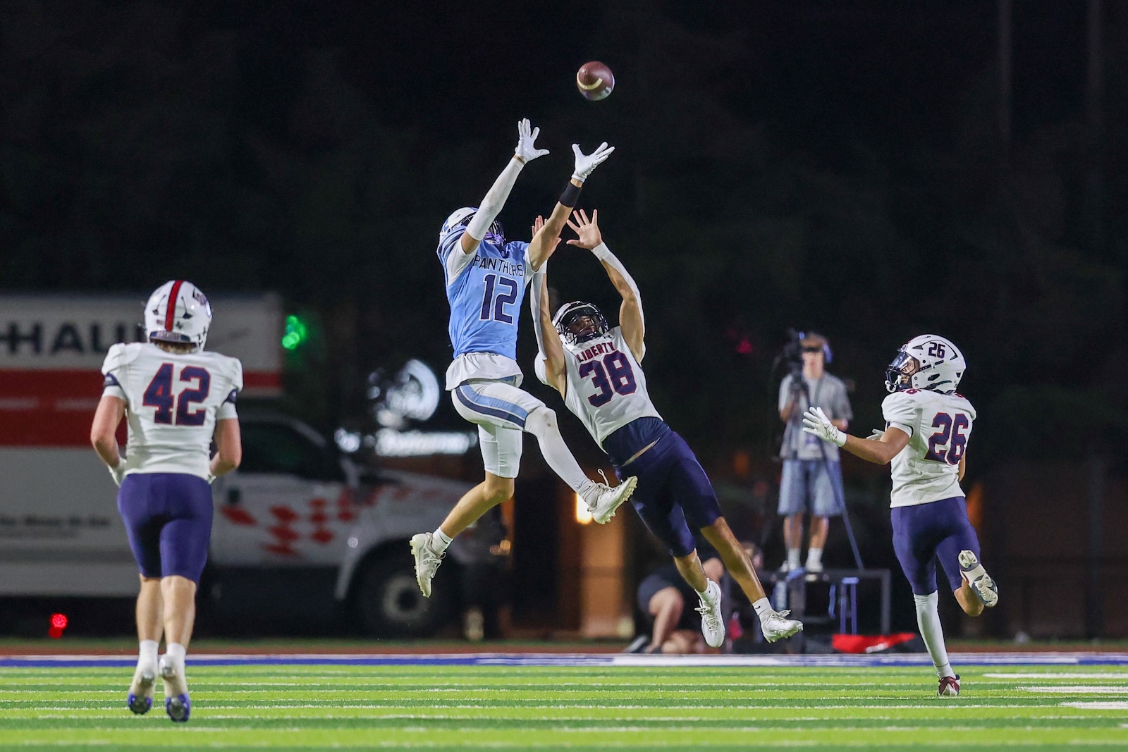 Seabolt Chase-16177 | Press Pass Sports Two football players leap to catch a lofted ball at night, with teammates nearby on a bright green field.