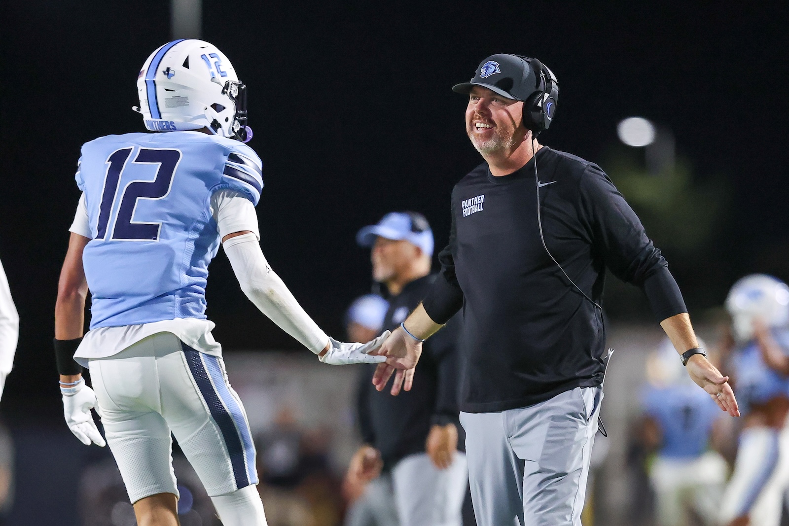 Seabolt Chase-16290 | Press Pass Sports Coach wearing a headset high-fives a football player in a light-blue uniform with the number 12 on a night field.