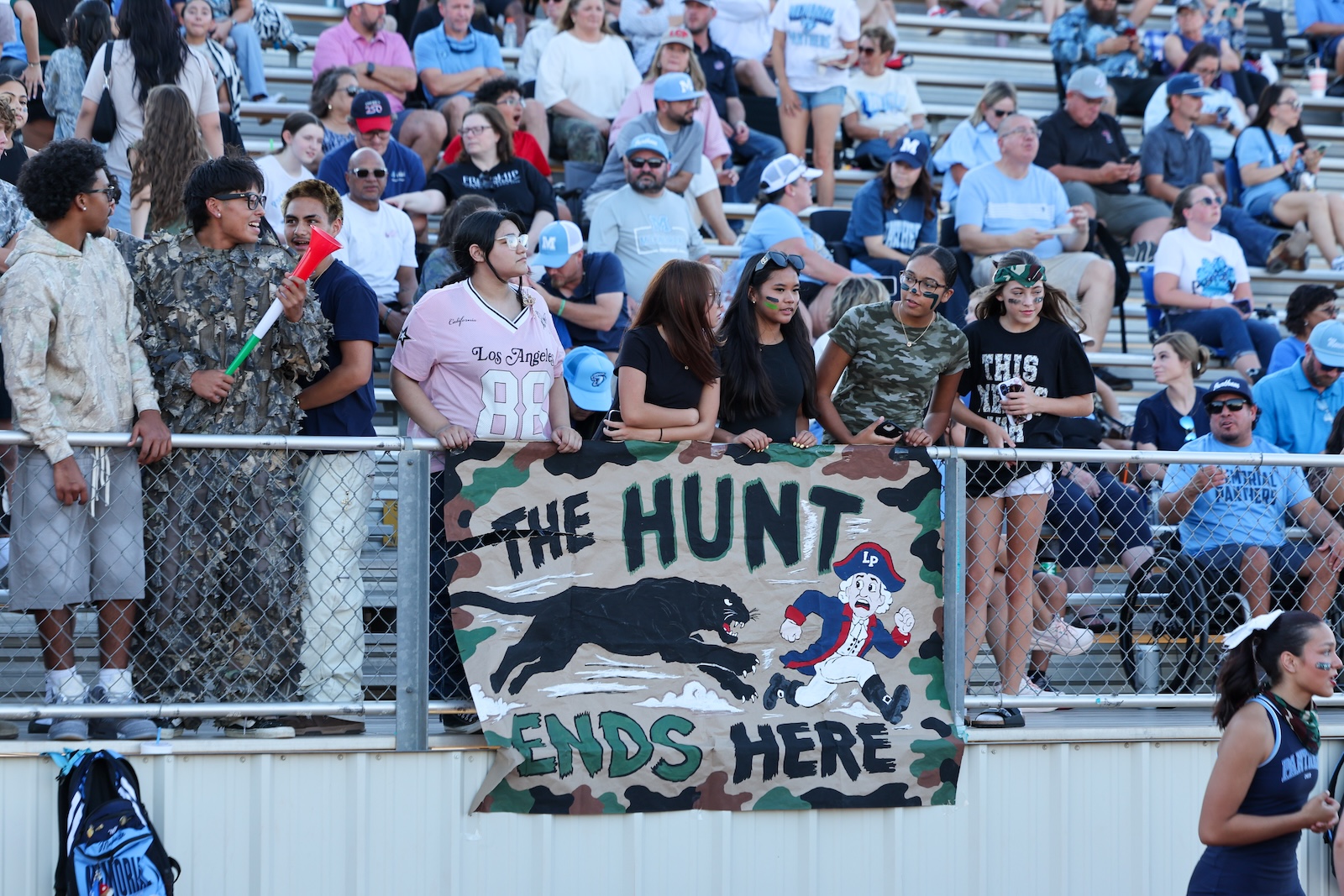 Seabolt Chase-24560 | Press Pass Sports Crowd of spectators at a stadium railing behind a camouflage banner that reads 'THE HUNT ENDS HERE' with a black panther and a cartoon pirate image.