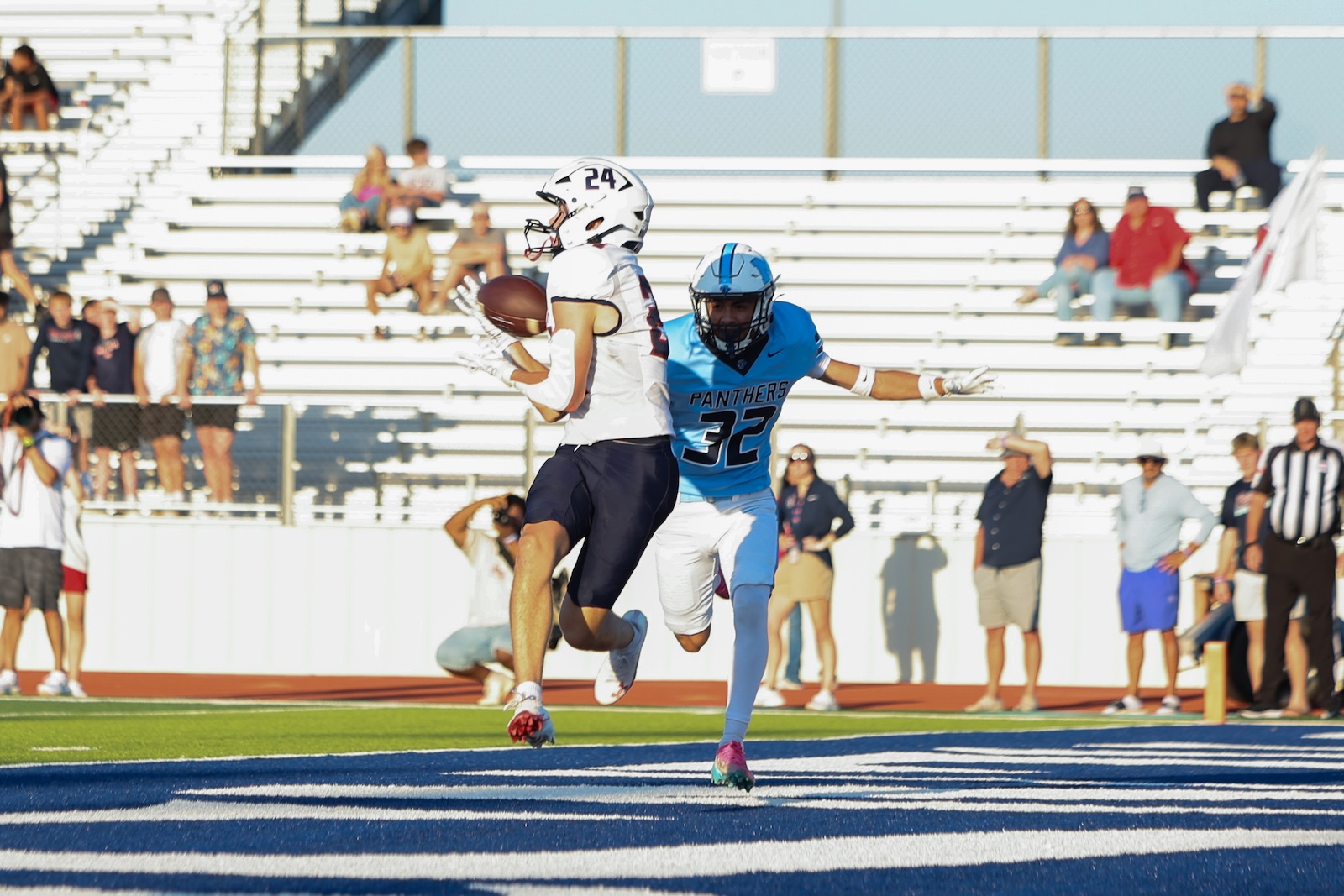Seabolt Chase-24573 | Press Pass Sports Football game: white-uniform receiver jumps with ball while pursuing end zone, pursued by a blue Panthers defender on a blue end zone turf, spectators in stands in background.