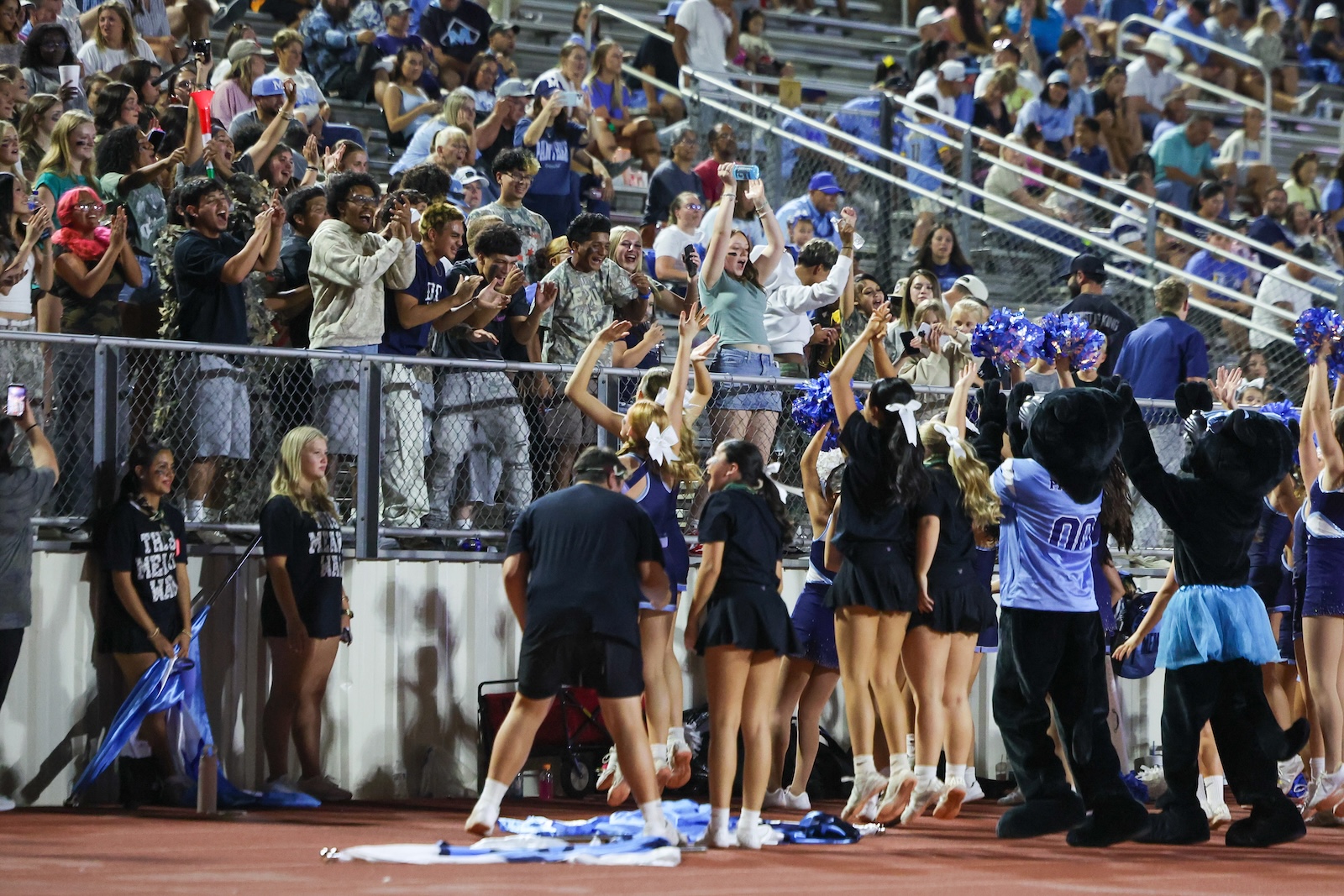 Seabolt Chase-24777 | Press Pass Sports Cheerleaders on the sideline facing a cheering crowd behind a chain-link fence at a stadium rallying with arms raised and pom-poms visible.