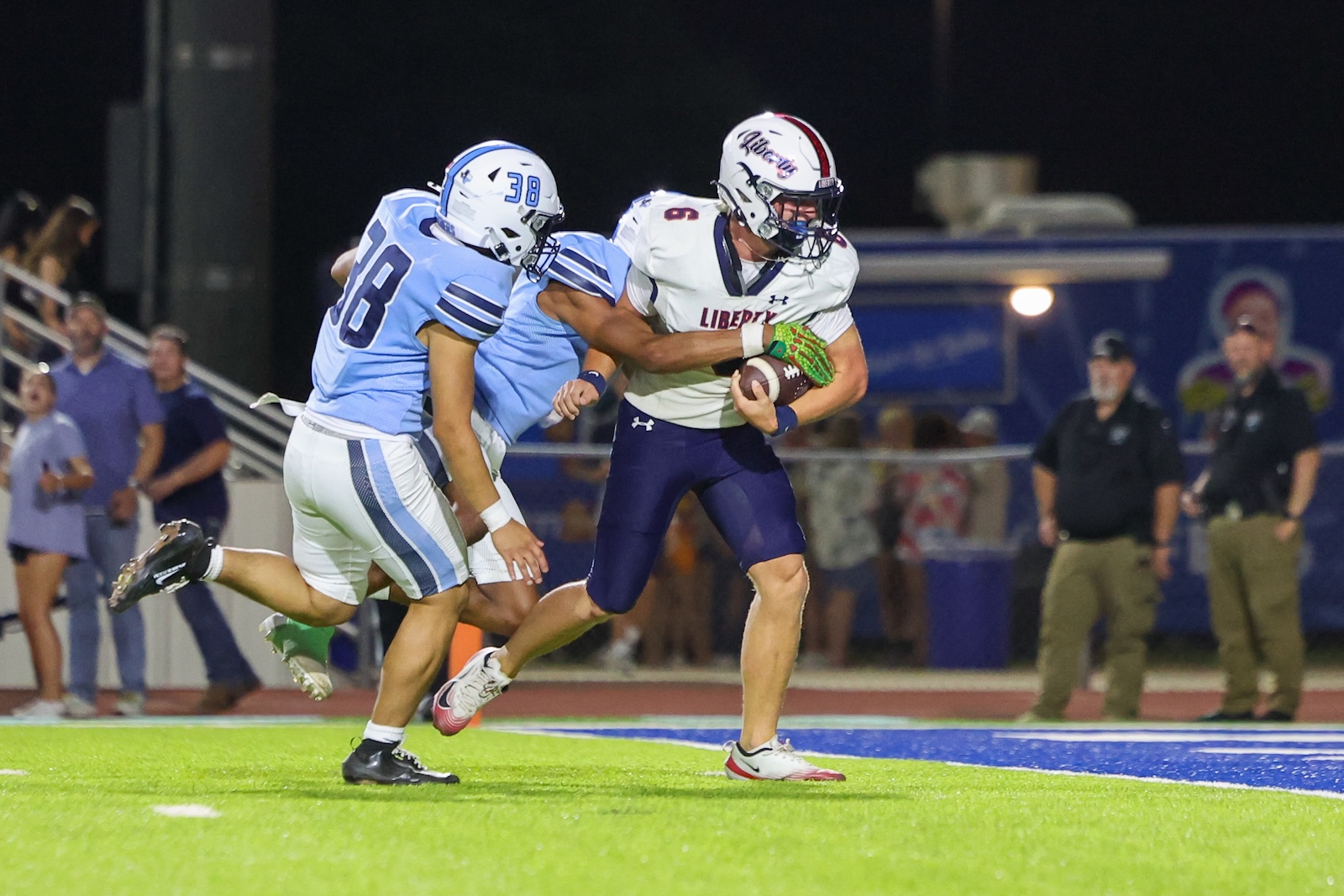 Seabolt Chase-24897 | Press Pass Sports Liberty football player in white jersey holds the ball while blue-clad defenders close in during a night game on a bright green field