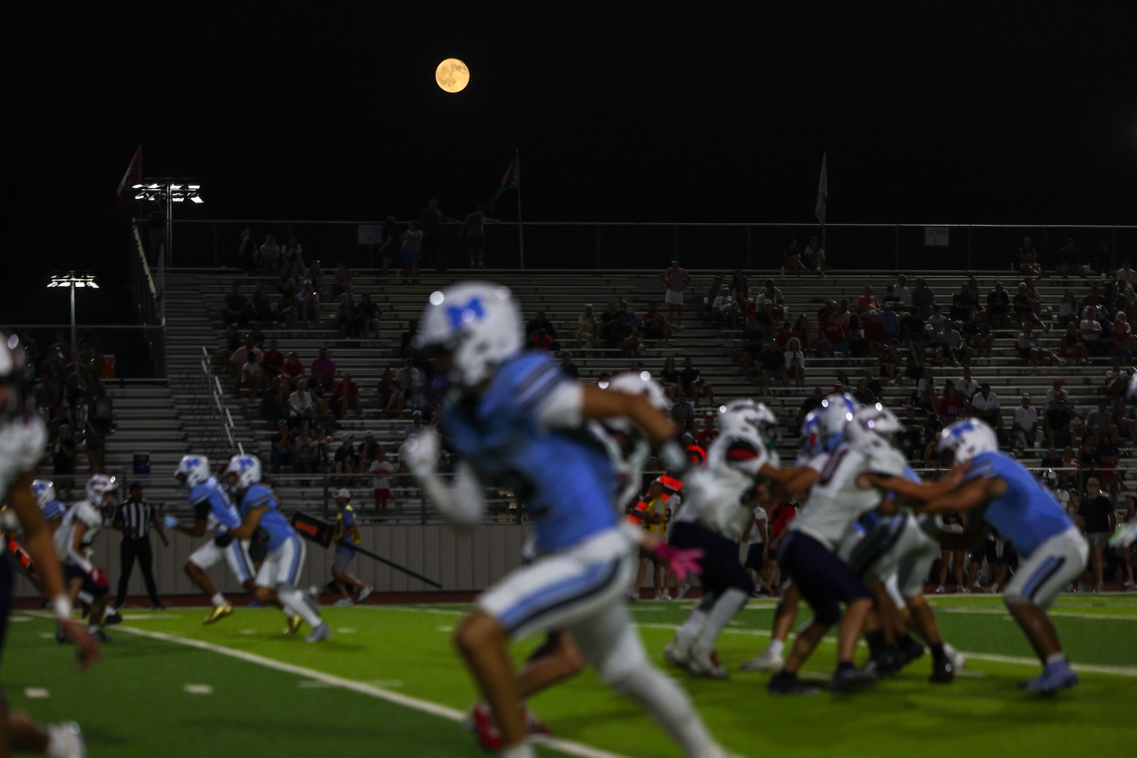 Seabolt Chase-25000 | Press Pass Sports Night football game on a bright green field; players in blue and white rush as a full moon rises over the stands with spectators.