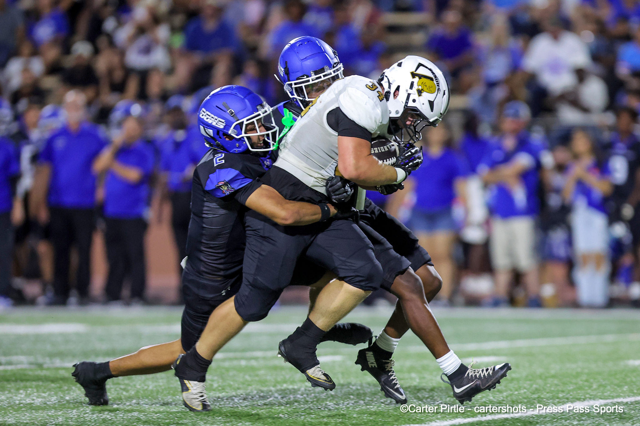 Two football players collide during a game: a blue-helmeted defender tackling a white-helmeted ball carrier on a green turf with a cheering crowd in the background.