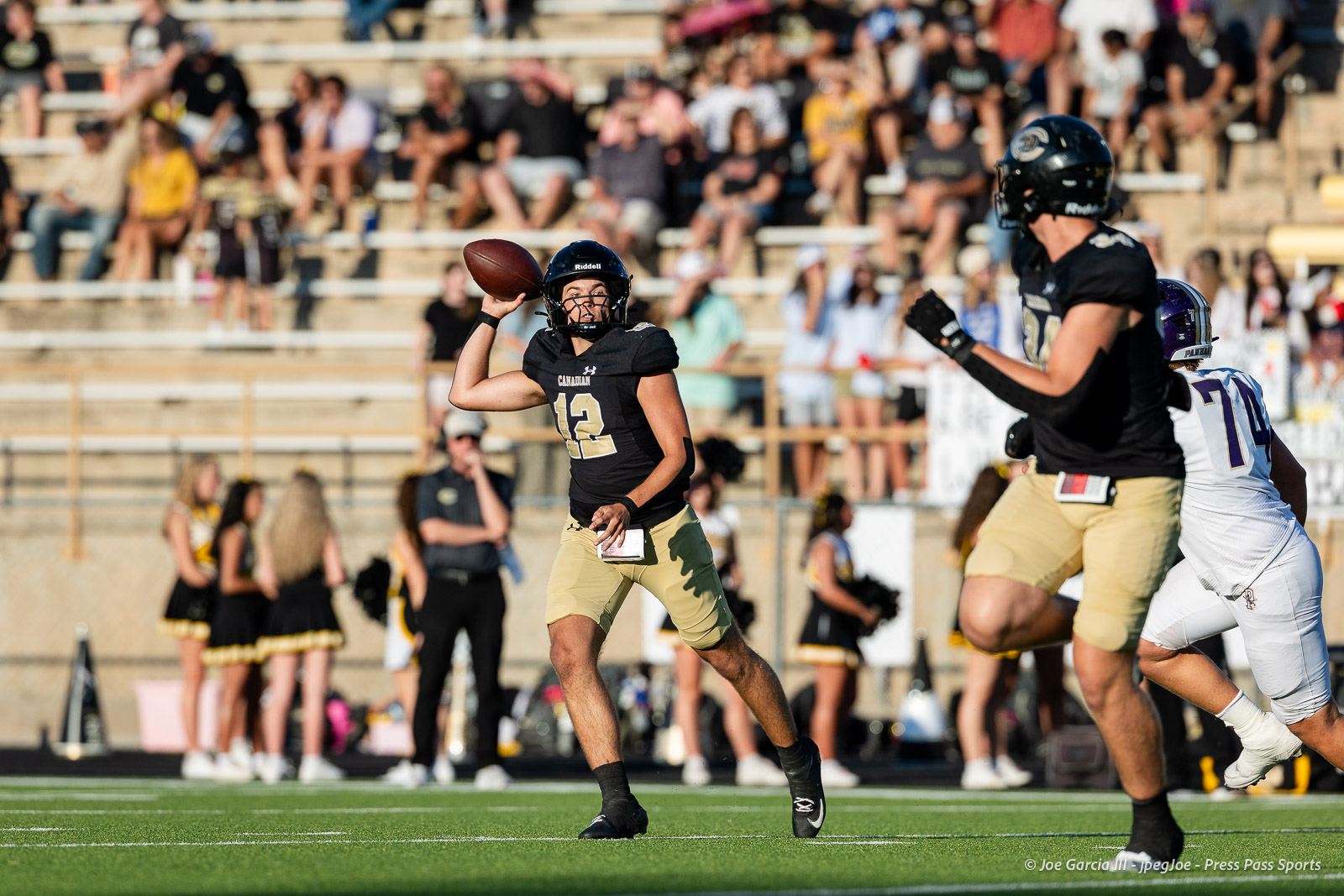 panhnandle-vs-canadian-football-jpegJoe-pps-3 | Press Pass Sports Quarterback in black and gold (number 12) throws a football on a sunlit field with a cheering crowd in the stands behind him.