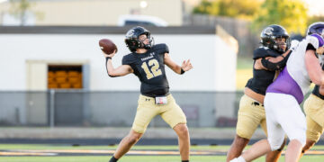 Quarterback in a black jersey (number 12) throws a football during a high school game while teammates block nearby on a green turf field.