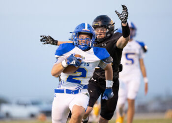 HHS-NHS-FB-52 | Press Pass Sports Football player in blue-and-white jersey runs with the ball while an opponent in black reaches from behind, on a sideline field.