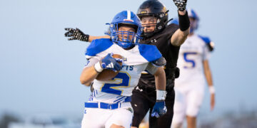 Football player in blue-and-white jersey runs with the ball while an opponent in black reaches from behind, on a sideline field.