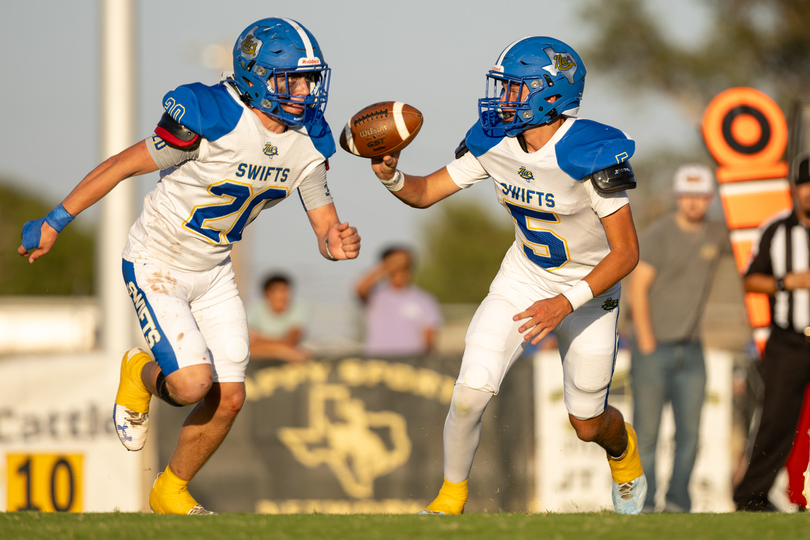 HHS-NHS-FB-32 | Press Pass Sports Two Swift players in white and blue uniforms reach for a football midair between them on a sunny field, both wearing blue helmets and pads, action shot at the sideline.