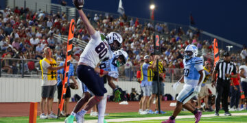 Player in white jersey reaches for a football over the goal line as teammates cheer in a blue end zone while a crowded stands watches at night.