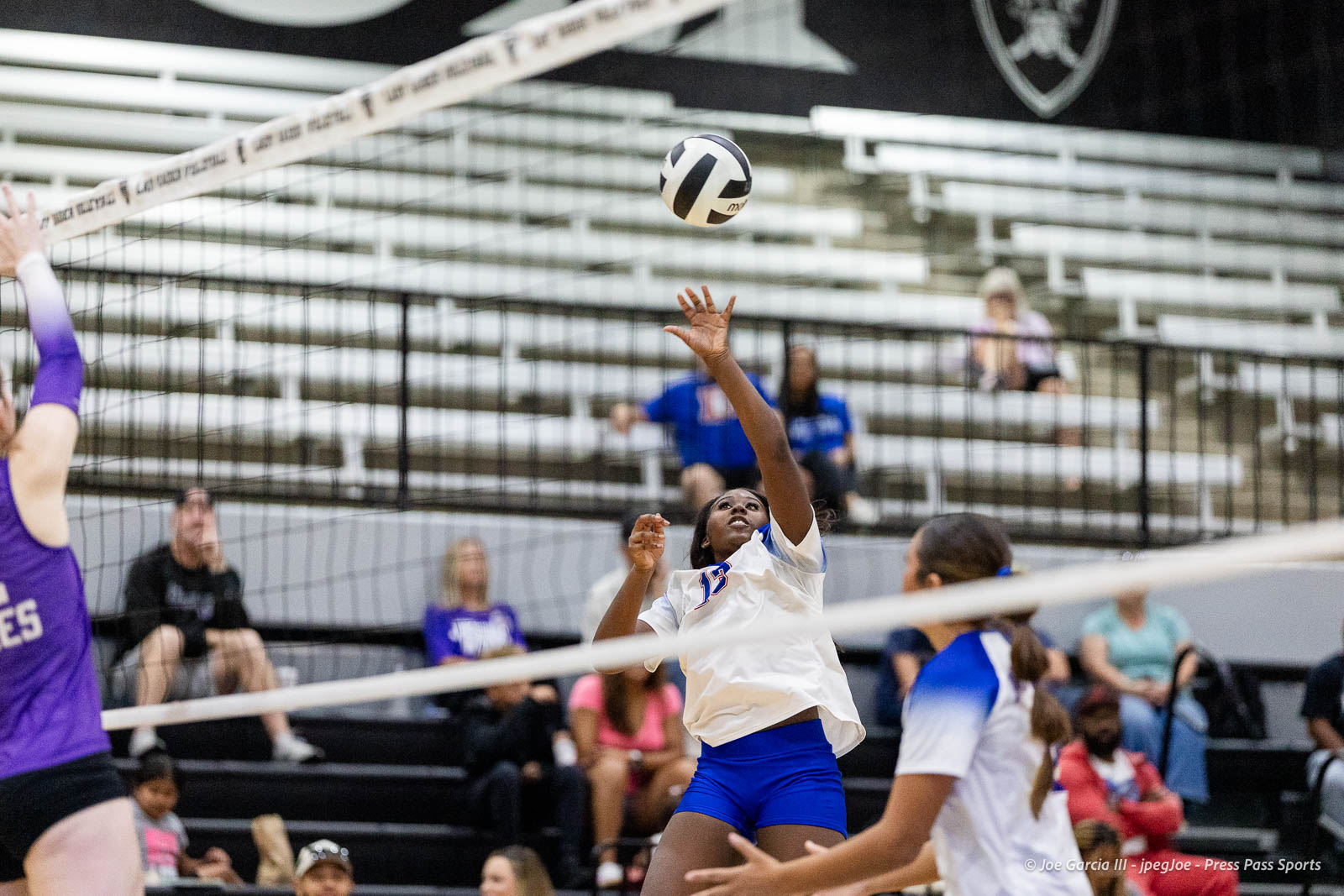 randall-showcase-2026-jpegJoe-pps-3 | Press Pass Sports Female volleyball player in white and blue reaching to hit a ball at the net during a match with spectators in the bleachers behind.