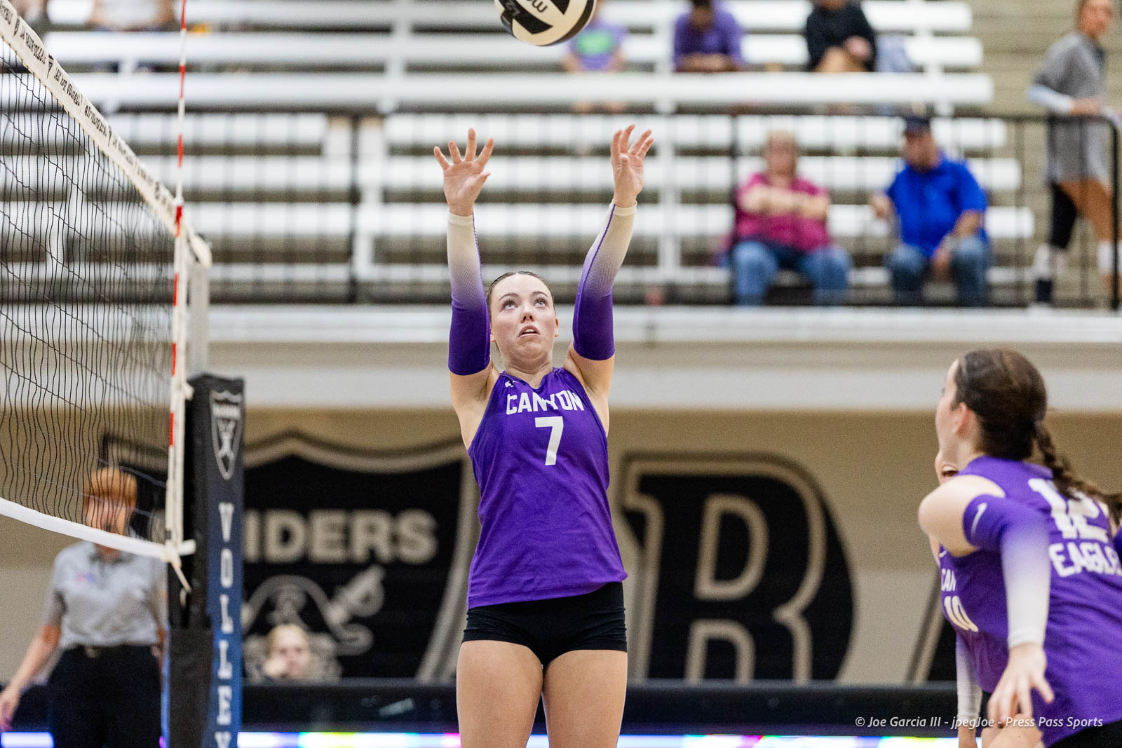 randall-showcase-2026-jpegJoe-pps-5 | Press Pass Sports Female volleyball player in a purple Canyon jersey (number 7) jumps with arms raised to contact the ball near the net in an indoor match. Background shows spectators in bleachers.