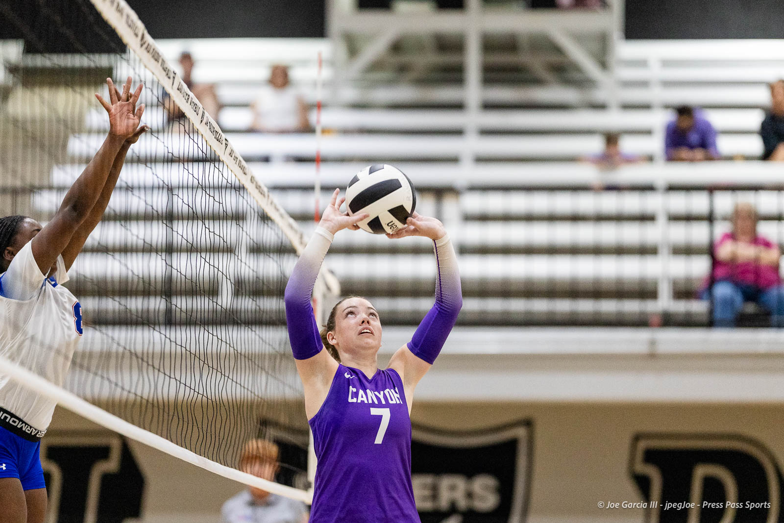 randall-showcase-2026-jpegJoe-pps-6 | Press Pass Sports A female volleyball player in a purple Canyon jersey #7 raises the ball for a set at the net while a defender jumps to block; gym bleachers in the background.
