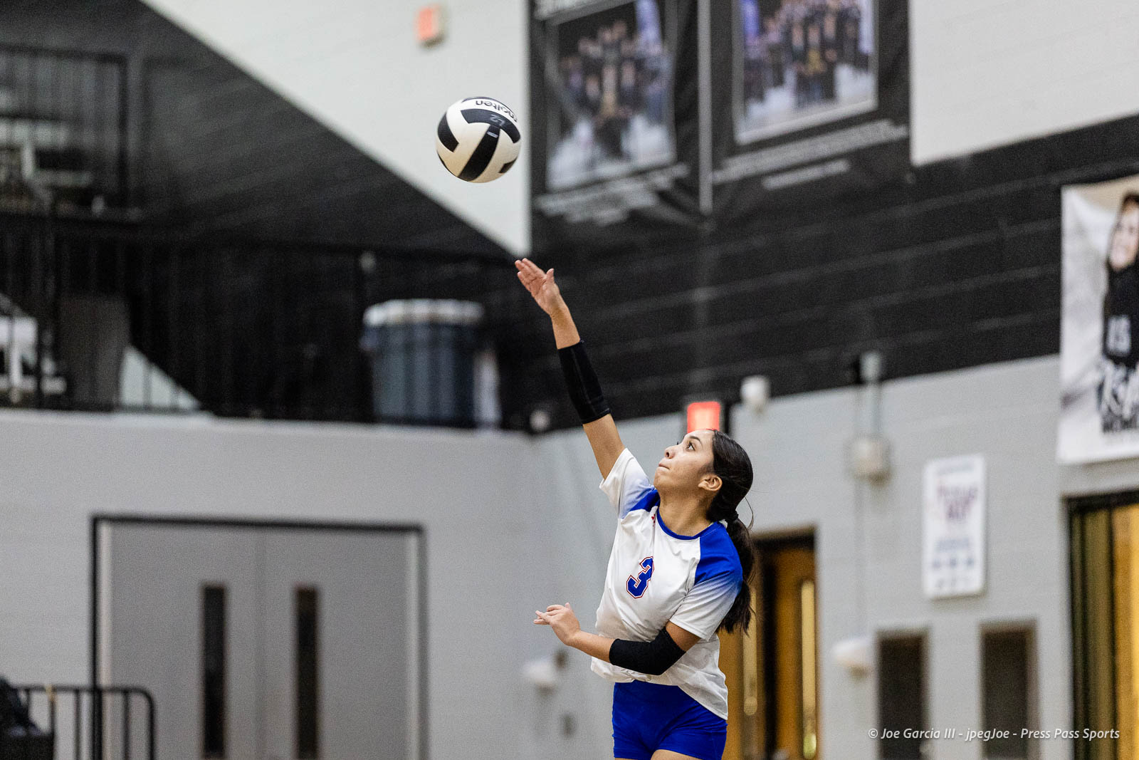 randall-showcase-2026-jpegJoe-pps-9 | Press Pass Sports Female volleyball player in white-and-blue uniform serves the ball indoors, right arm extended overhead, gym background behind her.