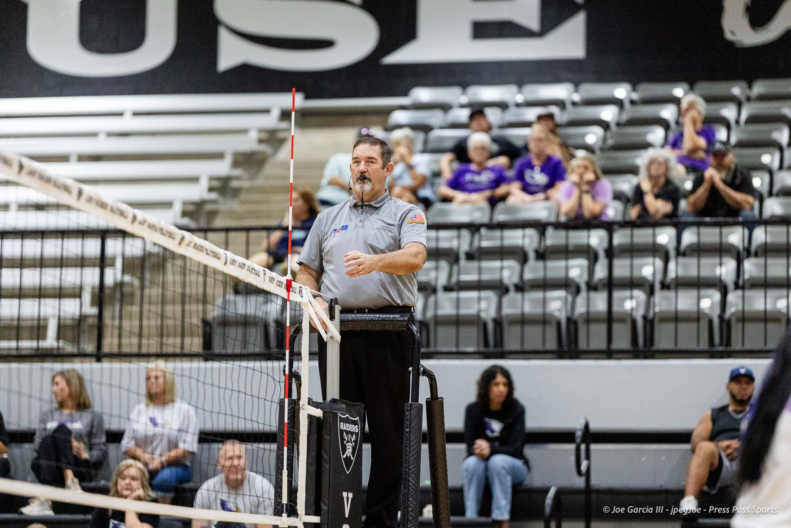 randall-showcase-2026-jpegJoe-pps-10 | Press Pass Sports Male volleyball referee in a gray shirt on a raised stand blowing a whistle beside the net, with spectators in the bleachers behind him.