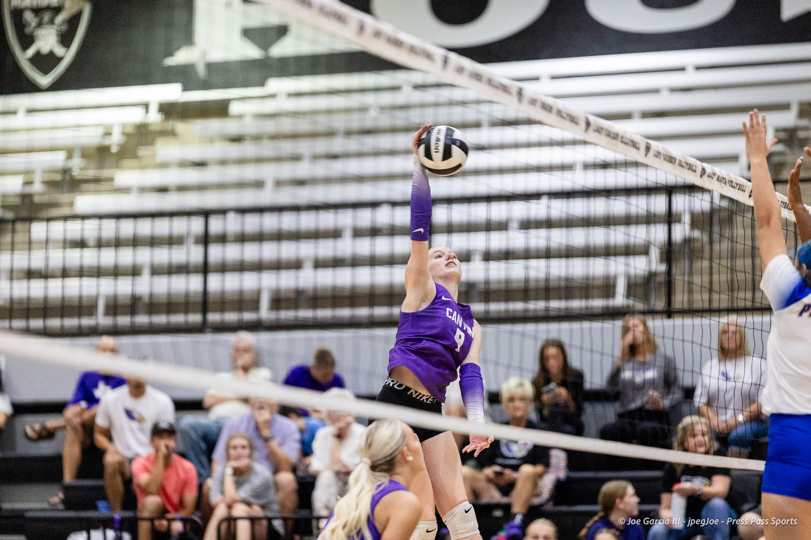 randall-showcase-2026-jpegJoe-pps-13 | Press Pass Sports Female volleyball player in a purple jersey jumps to spike the ball over the net during a match, with spectators in the bleachers behind.