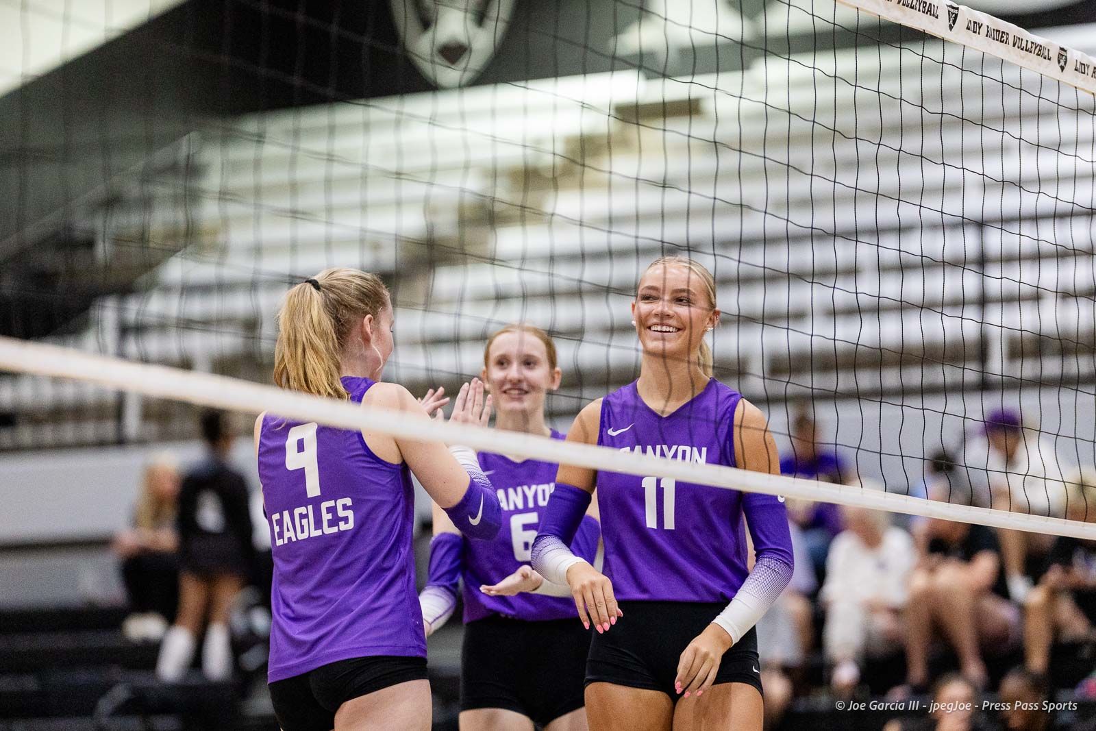 randall-showcase-2026-jpegJoe-pps-17 | Press Pass Sports Three female volleyball players in purple jerseys celebrate with high-fives at the net during a match.